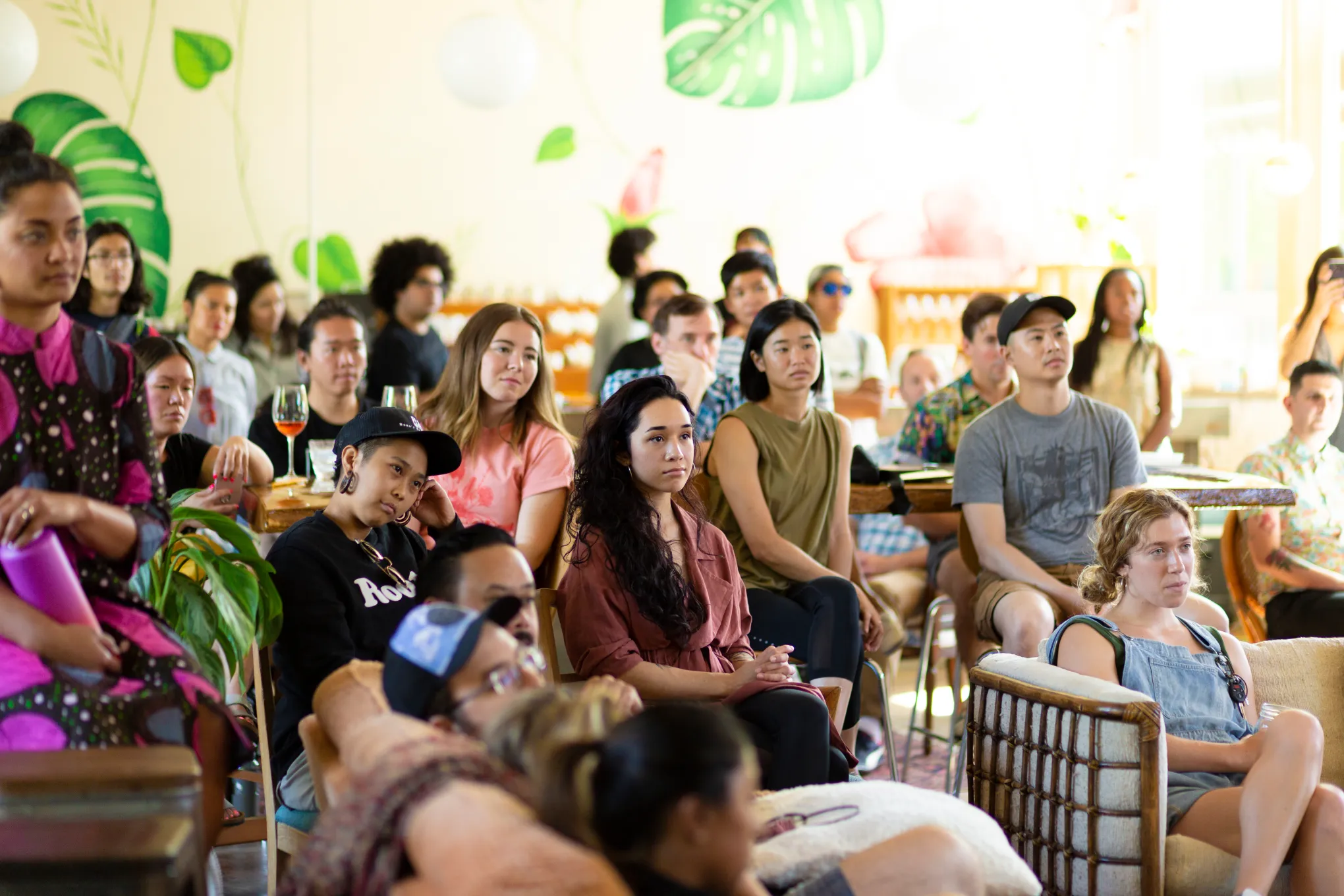Group of people seated indoors, some taking notes, with a bright, leafy background, attending a presentation or event.