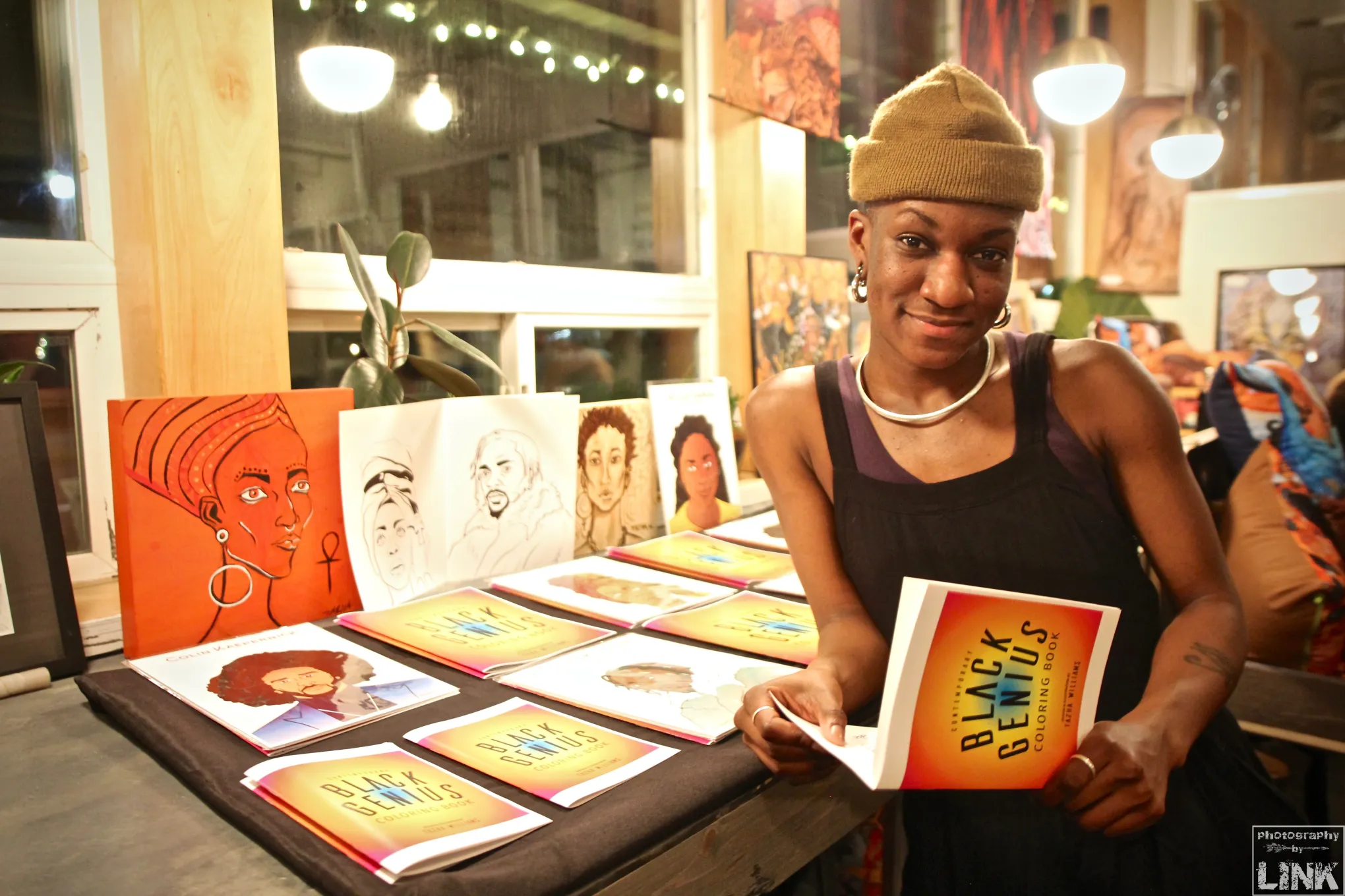 Woman in a black top and beige beanie holding a colorful book, standing near a table with artwork and books, inside a store.