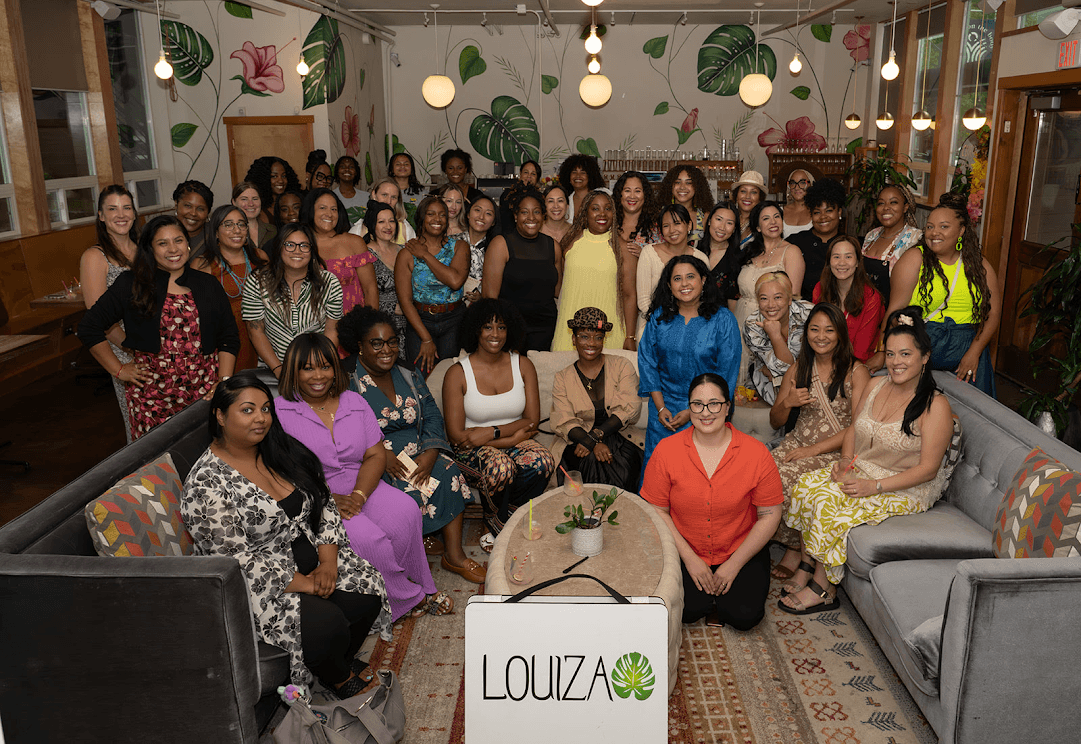 Group of women gathered in a decorated indoor space, some sitting and some standing, with plants and lights in the background.
