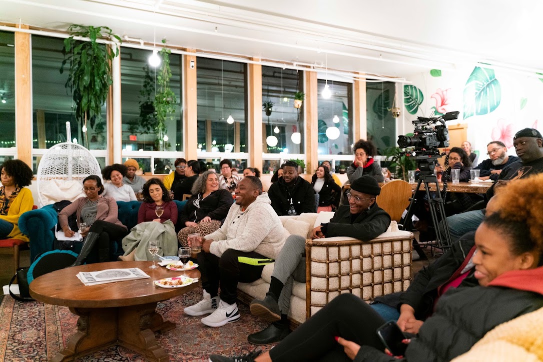 Group of people seated indoors, some watching a presentation or performance, with large windows and decorative lighting in the background.