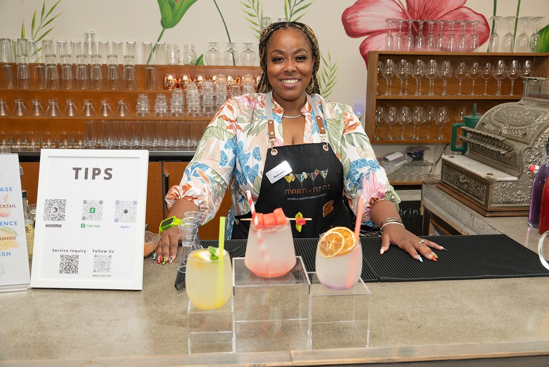 Smiling woman standing behind bar with colorful drinks, tips sign, and bar equipment, floral wallpaper background.