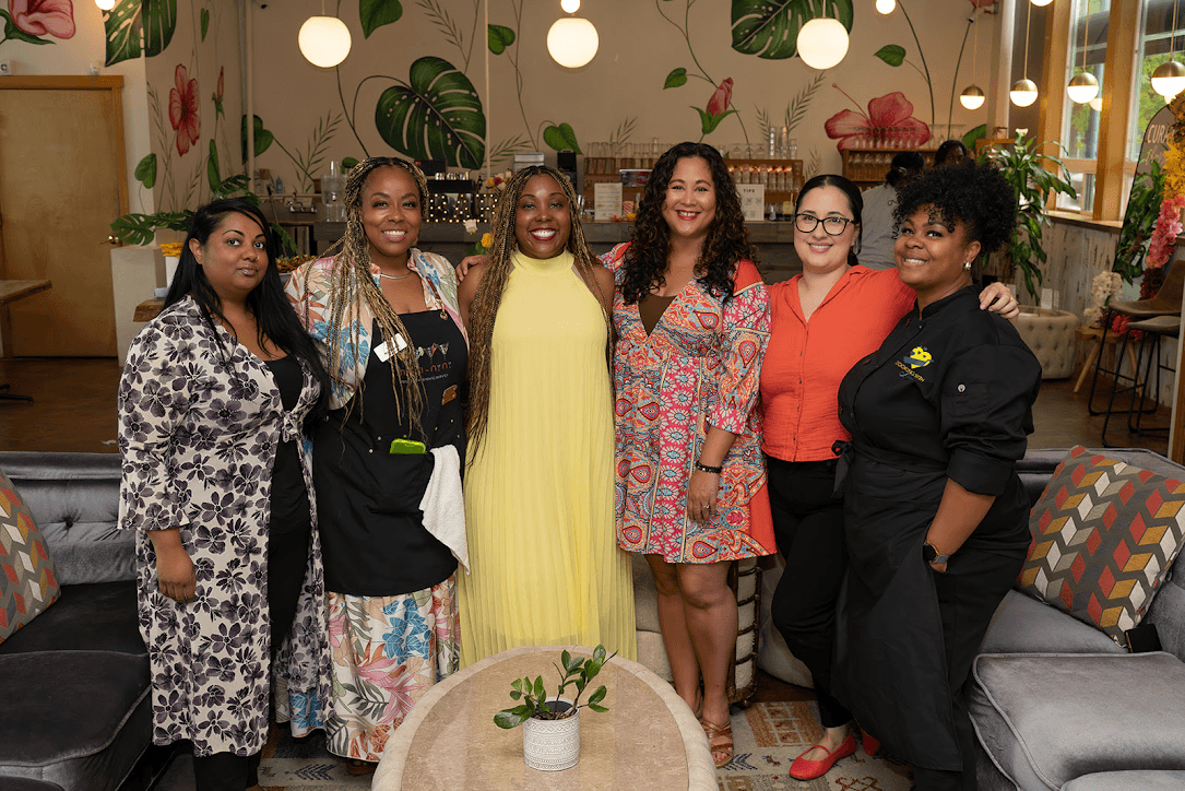 Six women standing together in a decorated indoor space, smiling, with plants and furniture in the background.