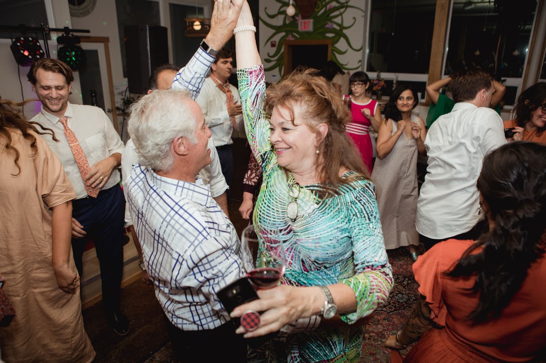 People dancing and smiling at a social gathering, with a woman holding a glass of wine and a man raising his hand.