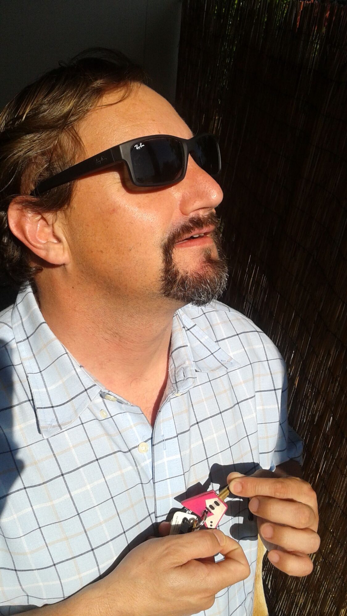 Man with sunglasses lying down, wearing a checkered shirt, holding a phone, with a dark background.