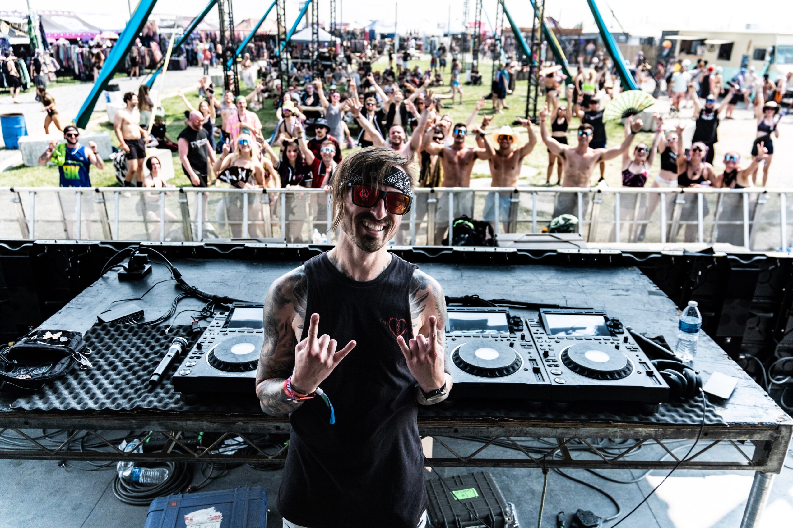 DJ performing at an outdoor music festival with a crowd dancing in the background, sunny day, DJ smiling, wearing black, hands raised.