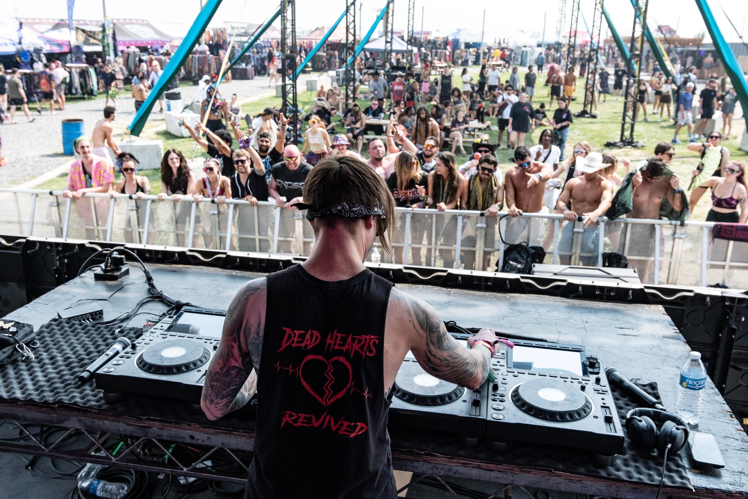 DJ performing at an outdoor festival with a crowd in the background, wearing a sleeveless shirt with a heart design.