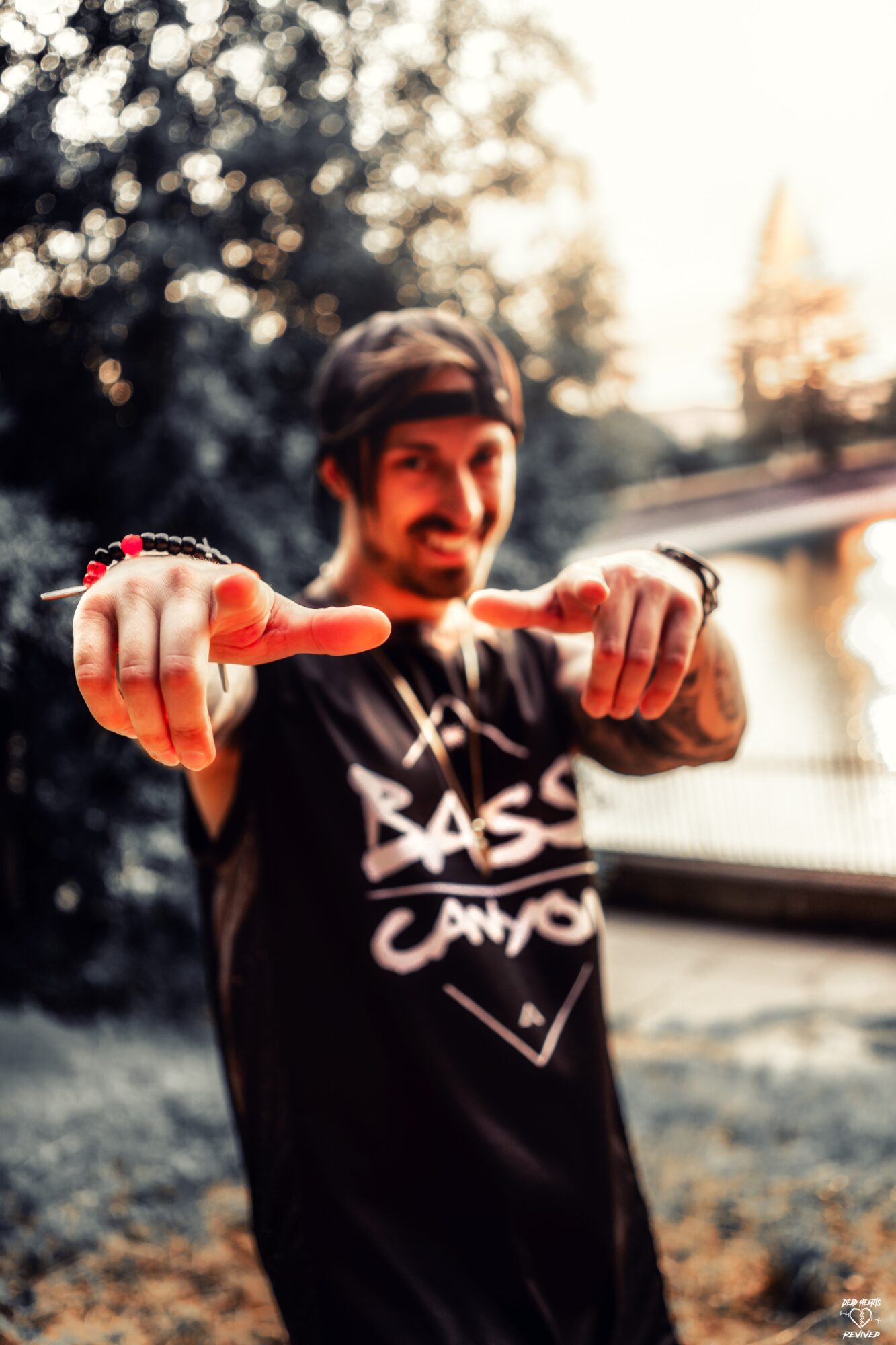 Person smiling outdoors, pointing towards the camera with both hands, trees and water in background