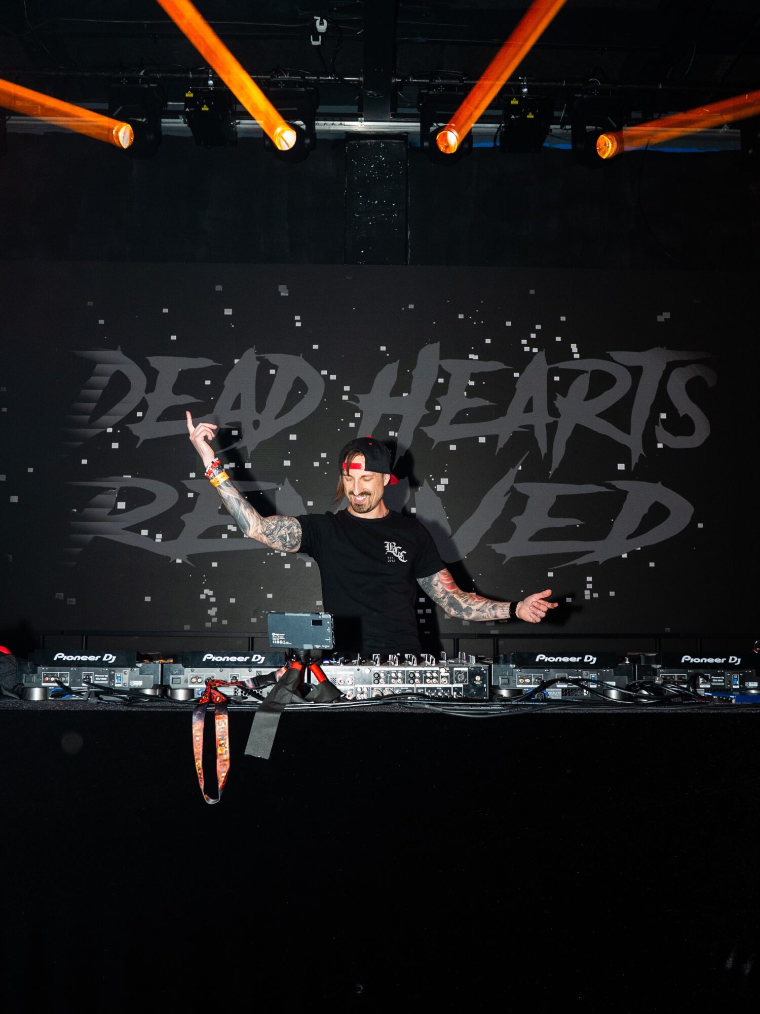 DJ performing on stage with orange lights overhead, behind a screen displaying 'DEAD HEARTS REMIXED', with DJ equipment in front.