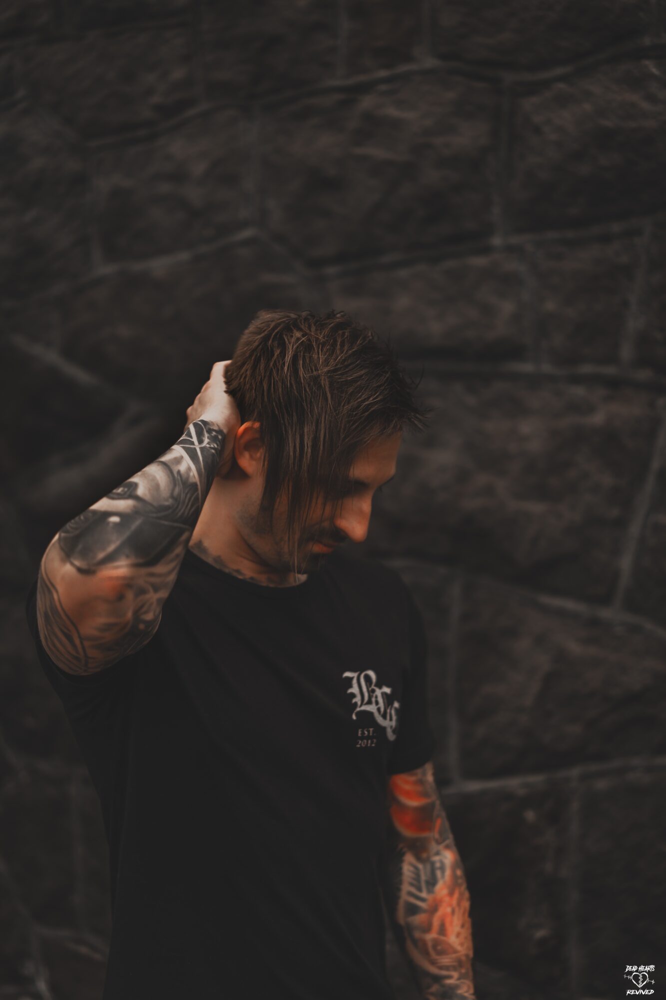 Young man with tattoos on arm, head tilted downward, hand behind head, dark background, wearing black shirt.