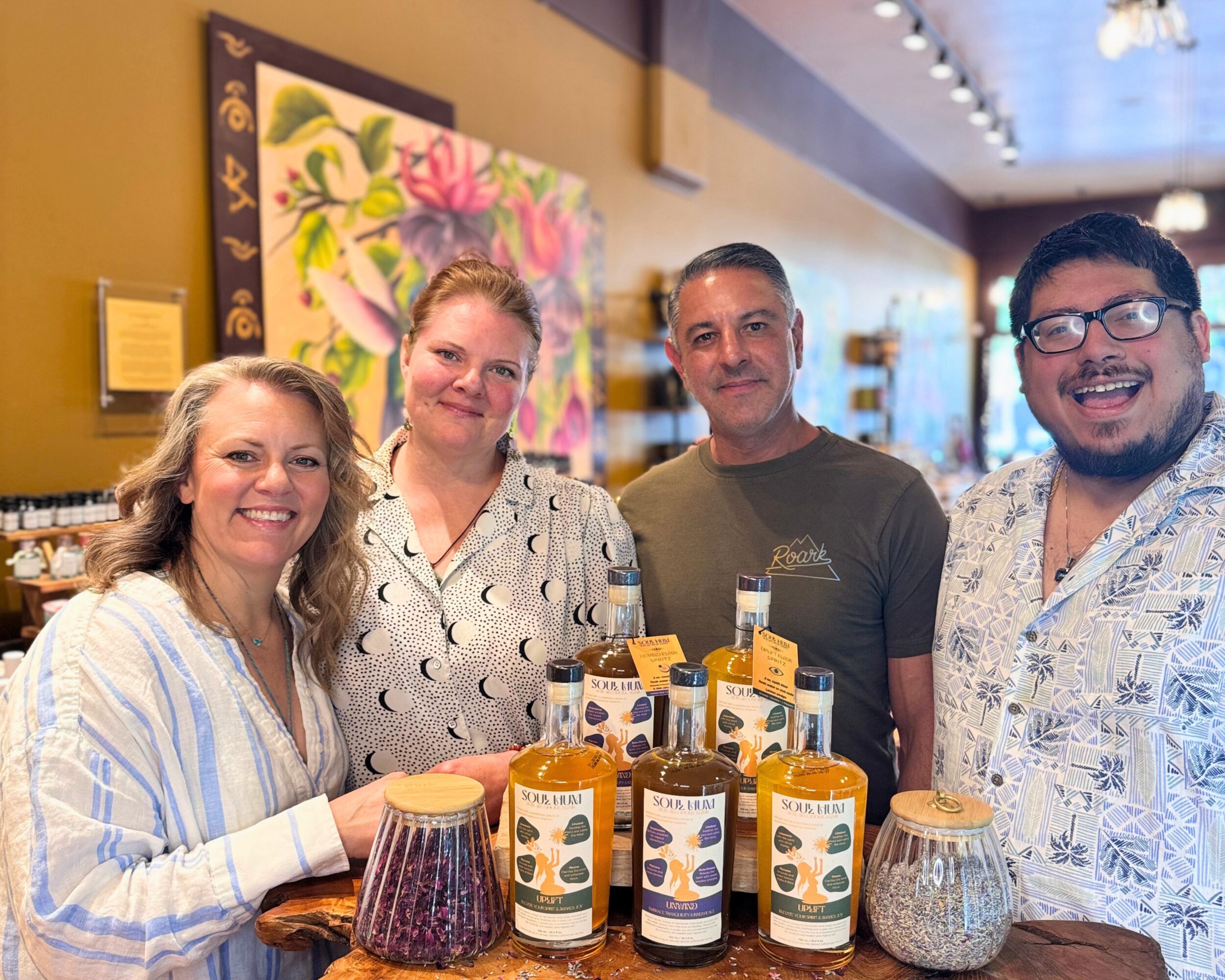 Four people smiling behind a table with jars and bottles, in a store or gallery setting.