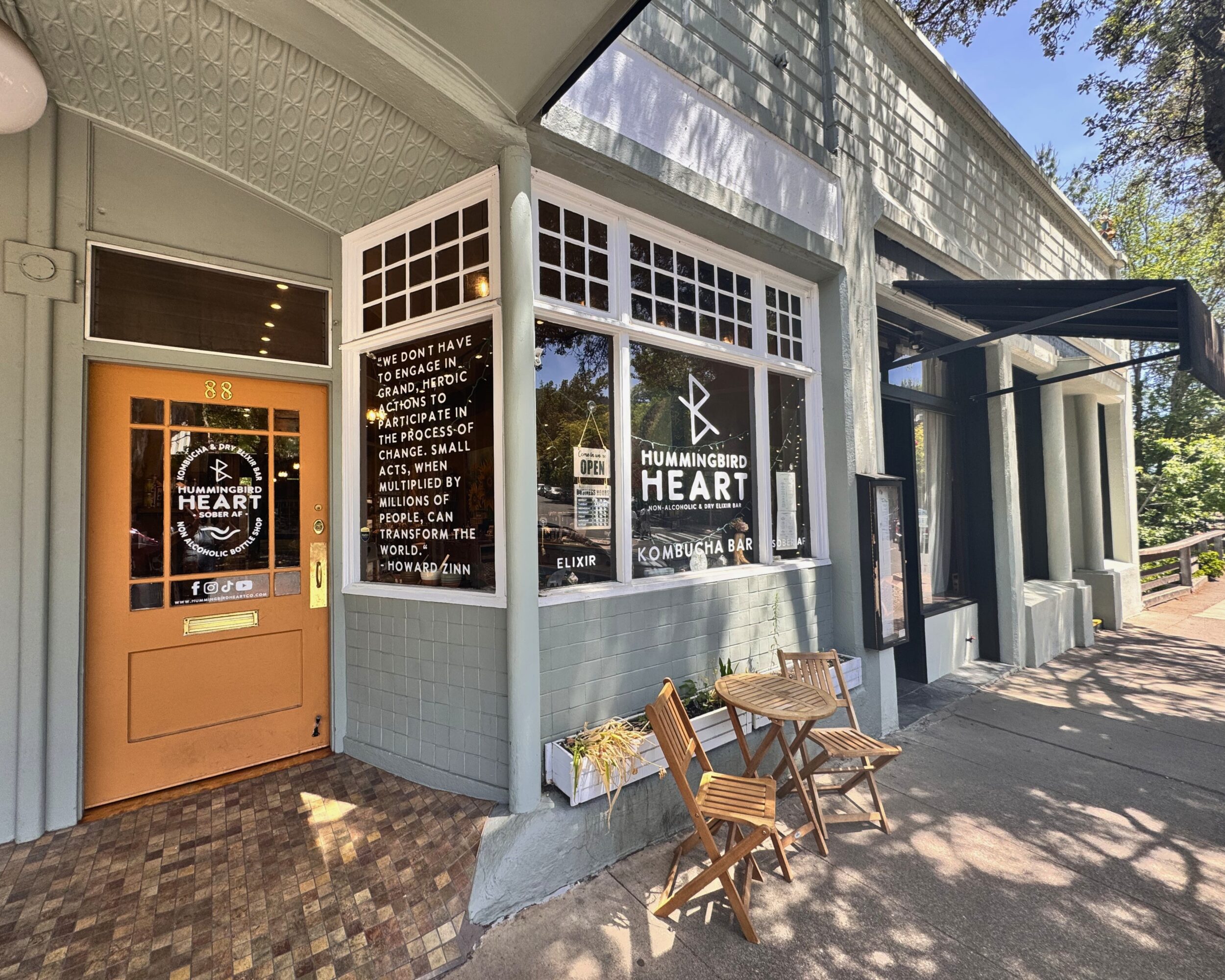 Storefront with large windows, two wooden chairs outside, and a door with a sign, under a shaded awning.