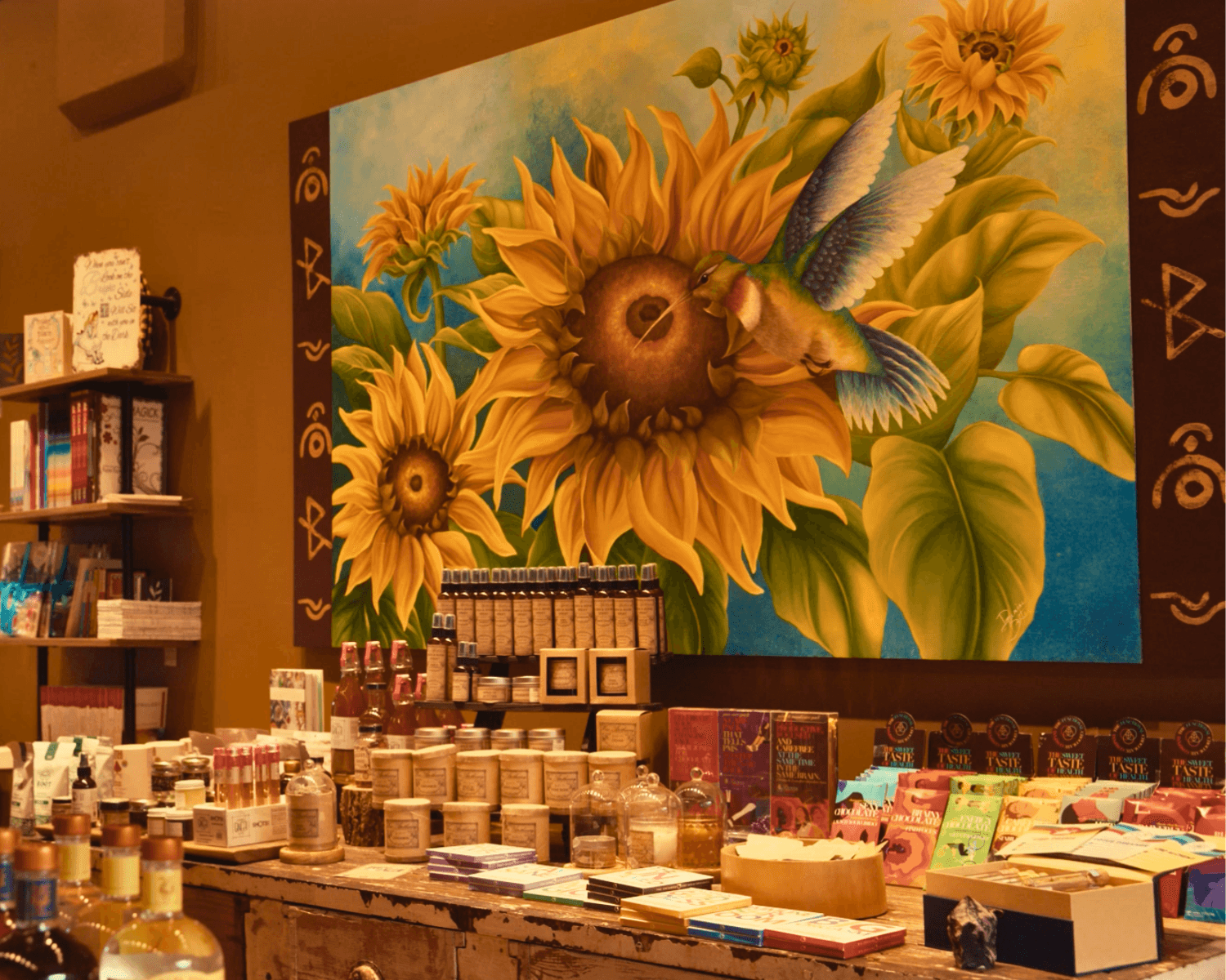 Colorful sunflower painting with a bird, displayed on a wall above a table with various products, in a store setting.