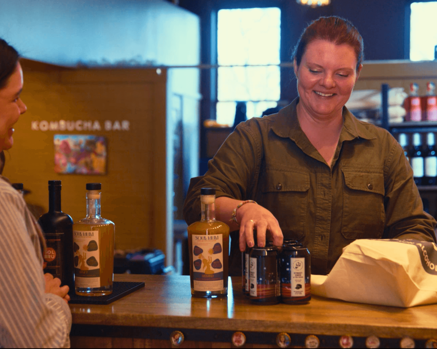 Woman smiling behind a bar counter with bottles and jars, engaging with a customer in a cozy setting.