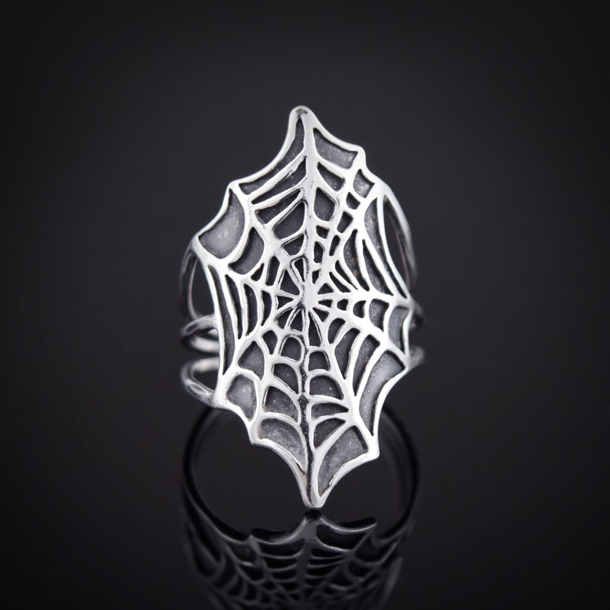 Ring with a spiderweb design on top, reflected on a dark surface, close-up, detailed, silver-colored, shiny, decorative.