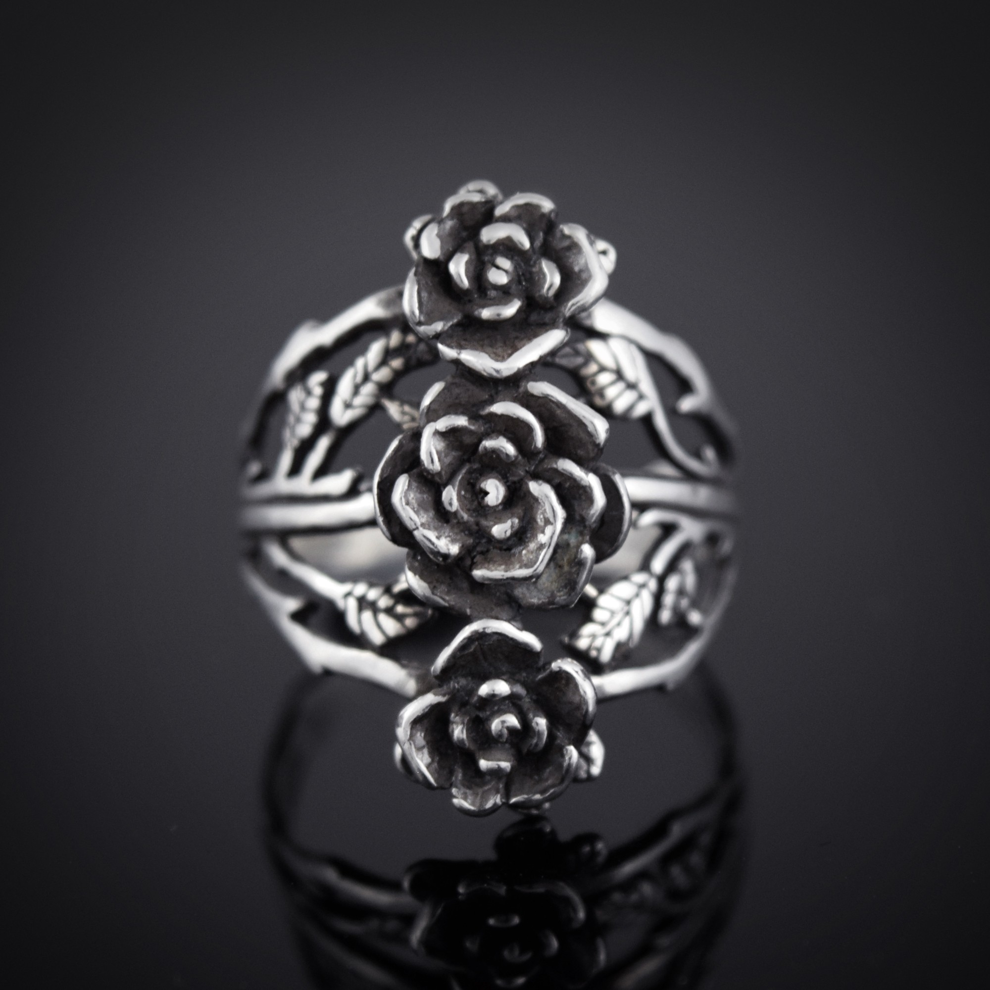 Silver ring with three detailed rose flowers and leaf patterns, reflected on a black surface.