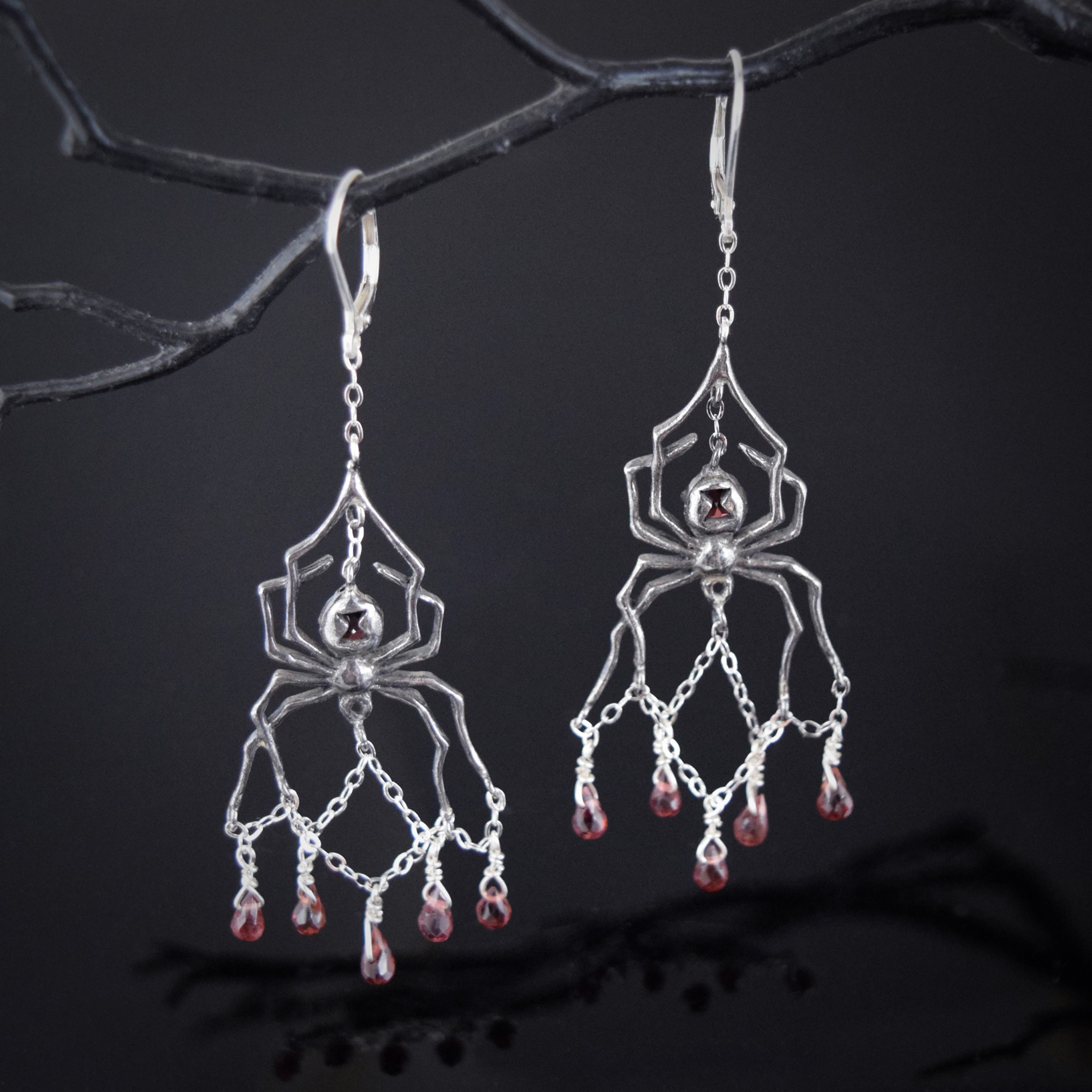 Silver spider-shaped earrings with red beads hanging from a black branch against a dark background.