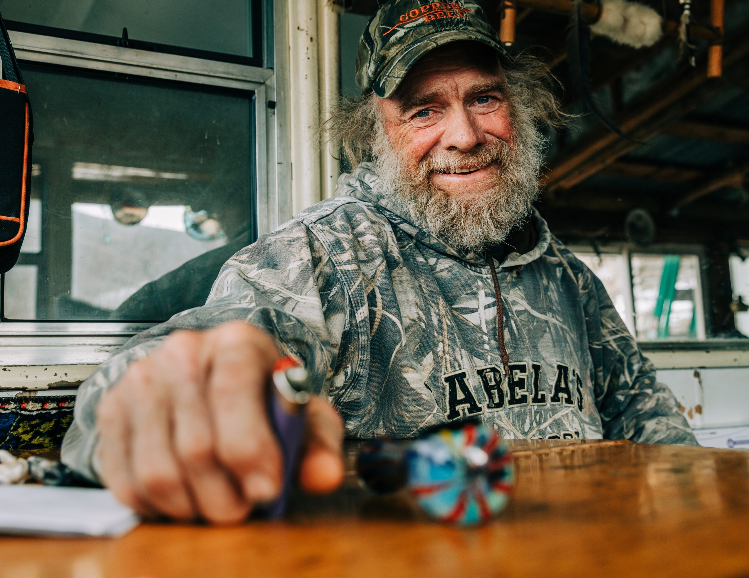 Smiling man with a beard wearing a camouflage hoodie and cap, reaching towards the camera, sitting at a wooden table.