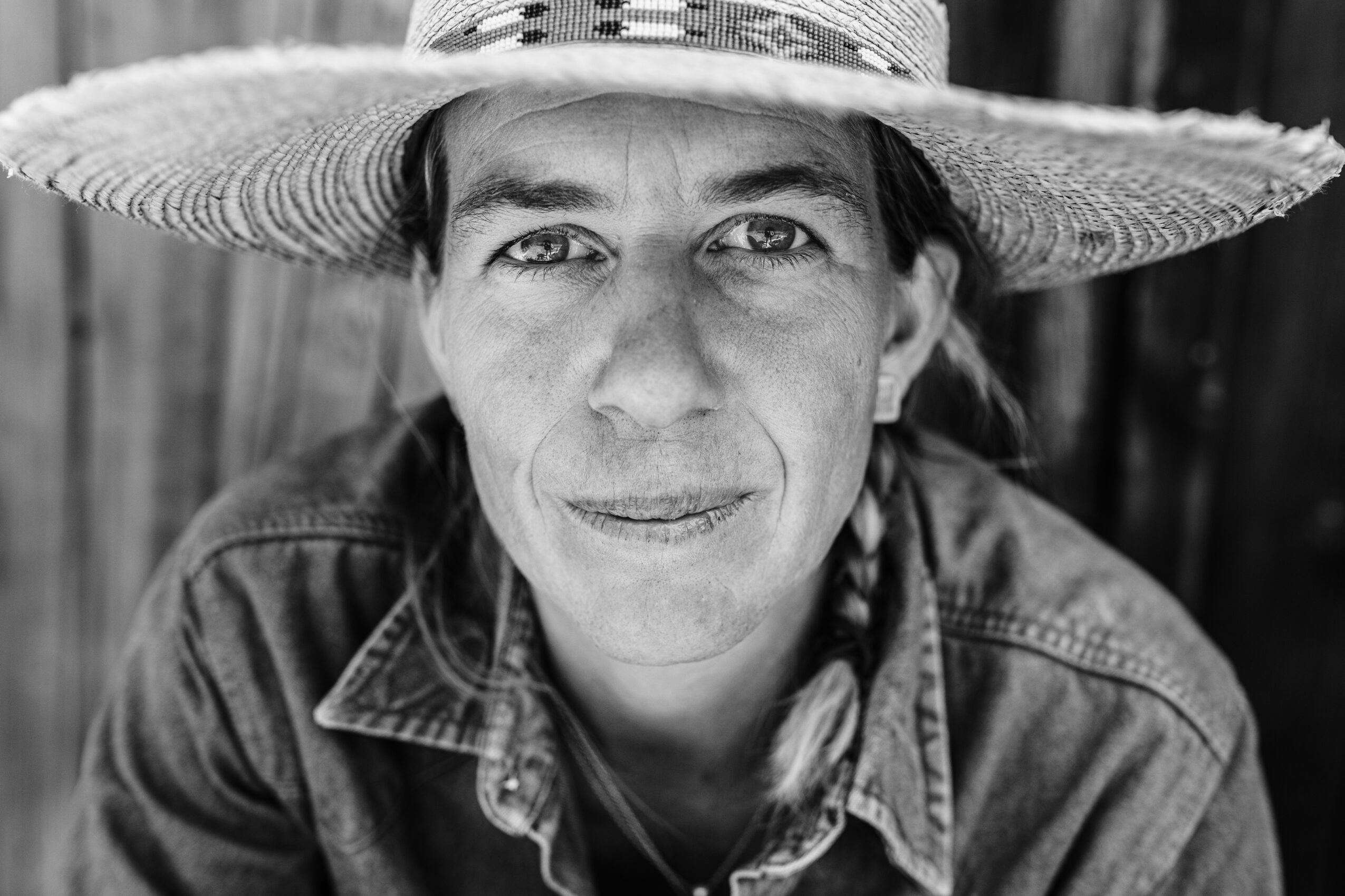 Woman wearing a wide-brimmed hat and denim shirt, smiling, outdoors with wooden background.