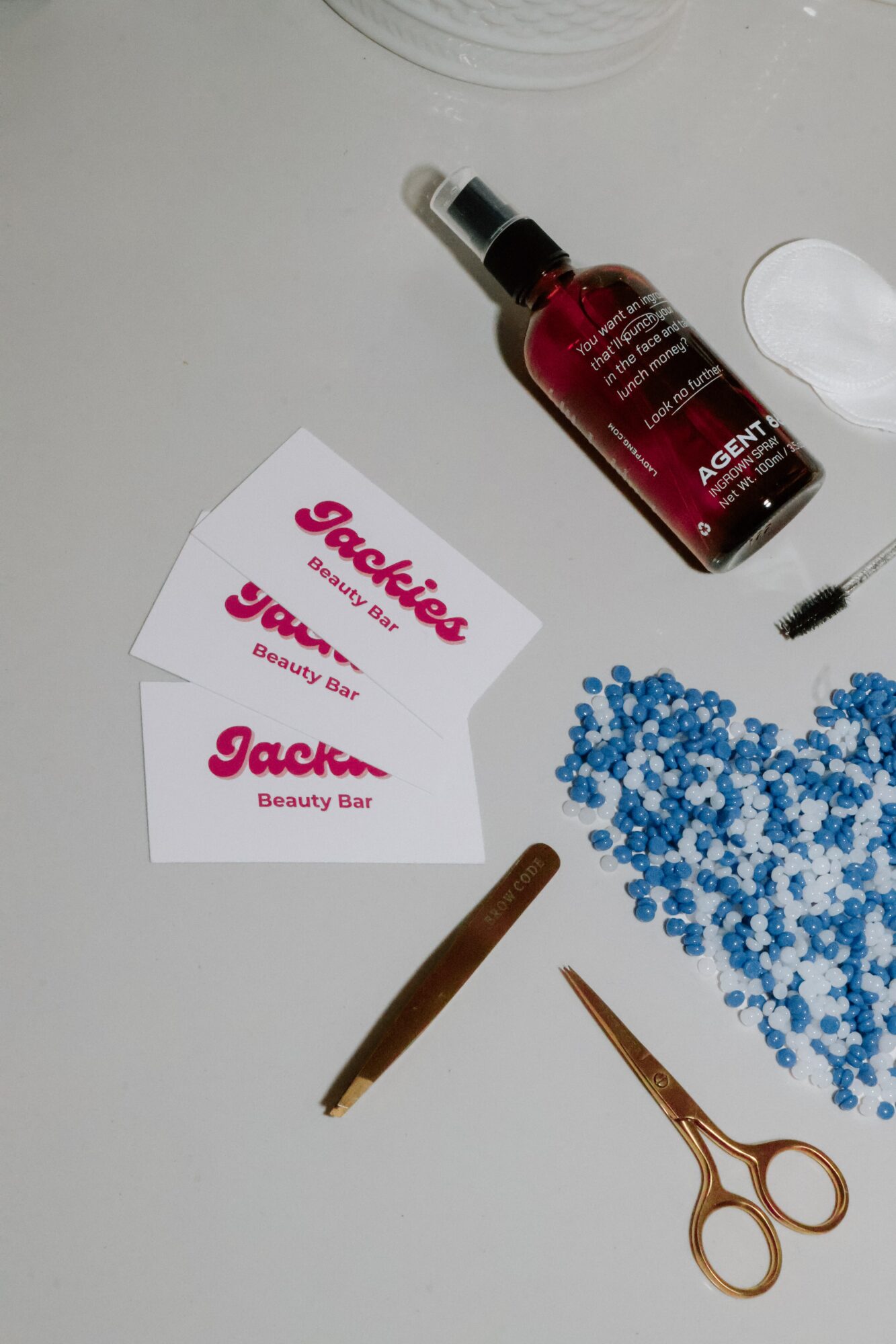 Two beauty bar cards, a bottle, white spoon, blue beads, pencil, and scissors on a light surface.