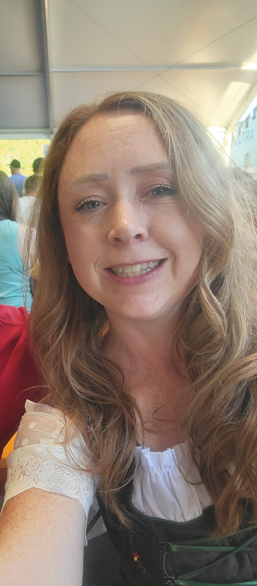 Woman with long wavy hair smiling, wearing a white top and red clothing, in a bright indoor setting.