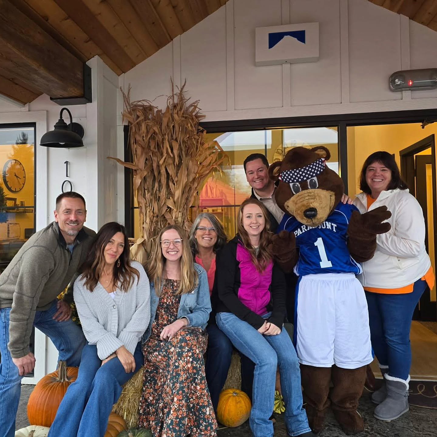 Group of six people and a bear mascot posing indoors with pumpkins, smiling, in a cozy setting.