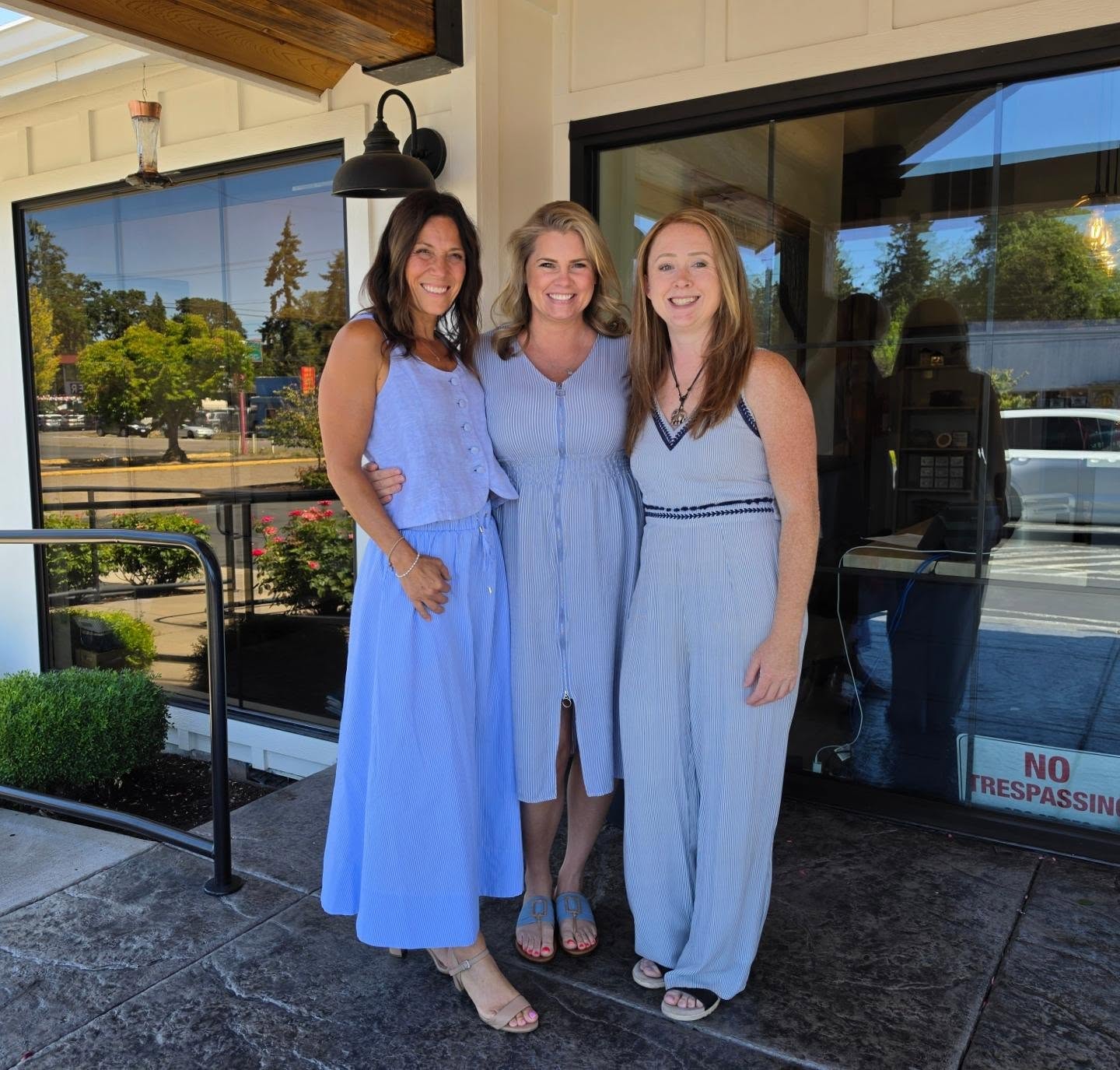 Three women standing outside near glass doors, smiling, with trees and cars in the background.
