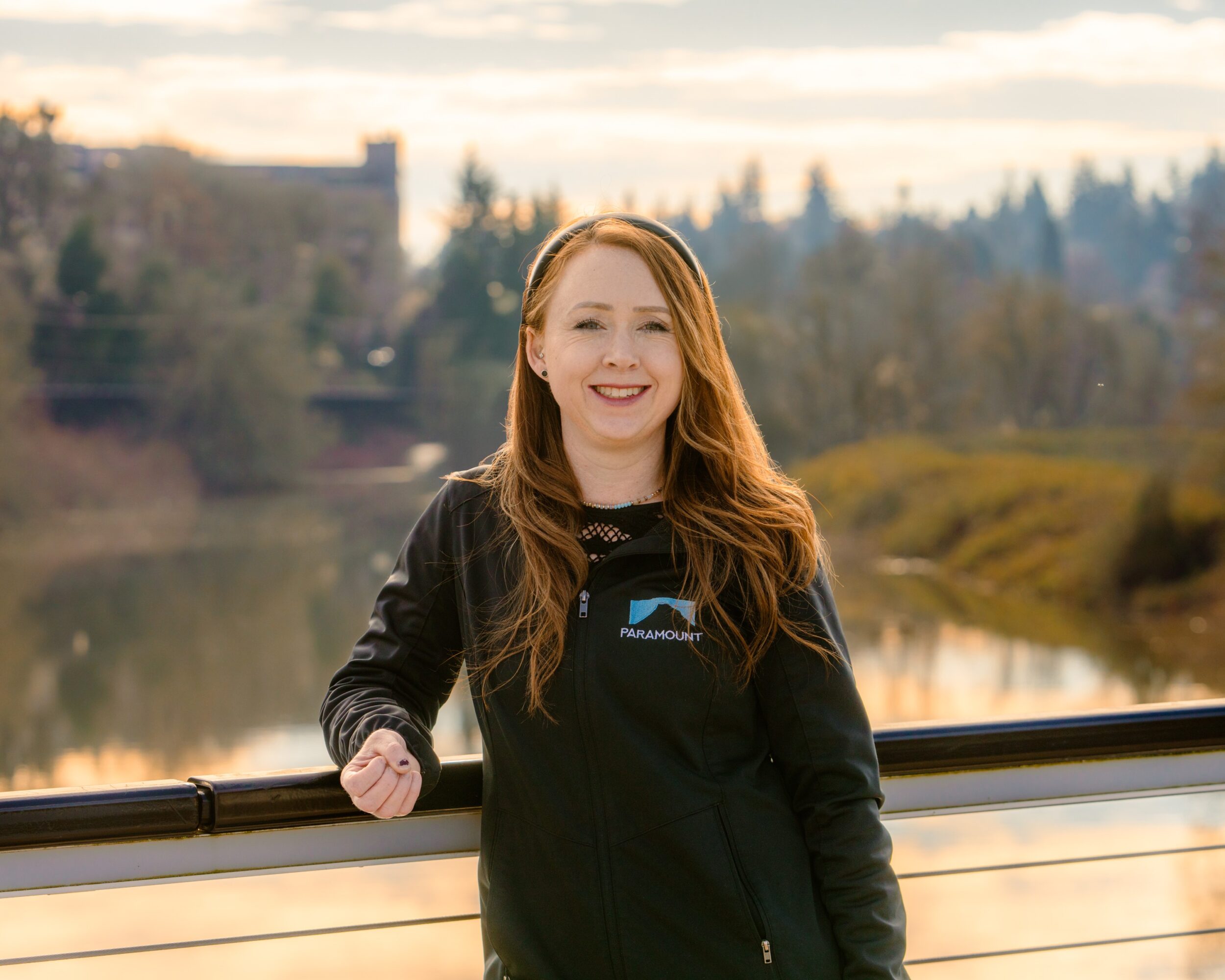 Young woman with long red hair smiling outdoors near a river, wearing a black jacket, during daytime.