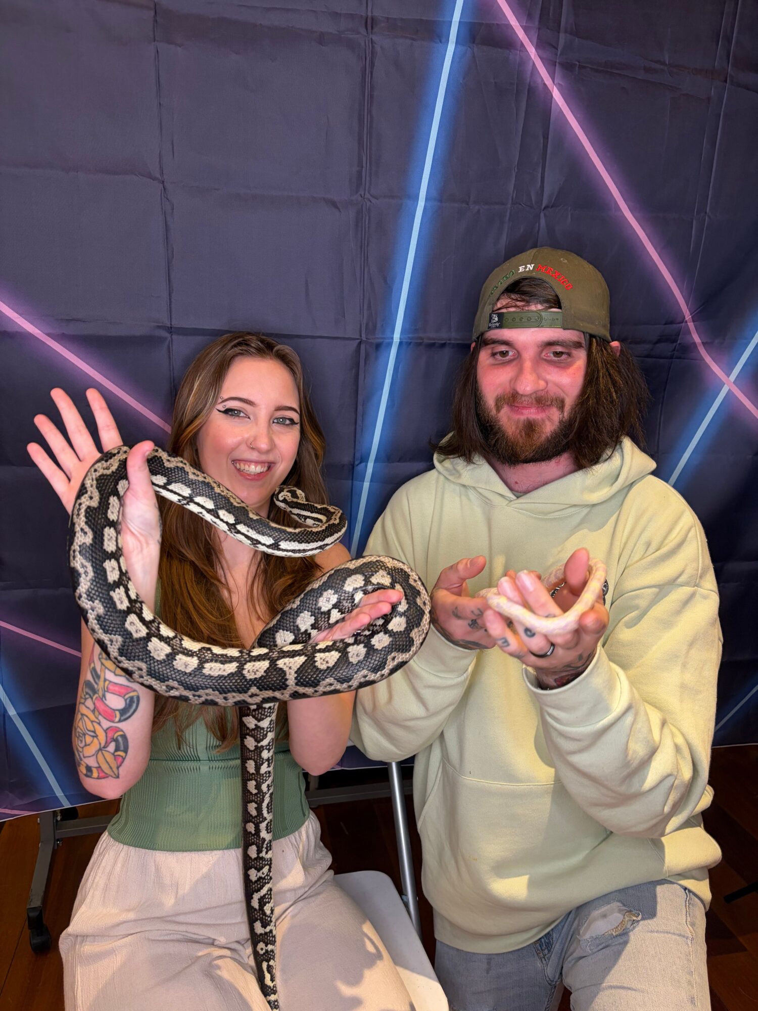 A man in a hoodie and cap holds a snake, with a woman beside him, both smiling, in front of a dark background with neon lights.