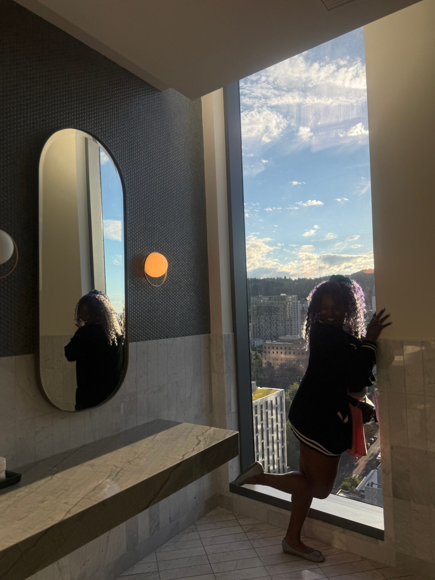 A person stands beside a floor-to-ceiling window in a modern bathroom, with a city view at sunset.