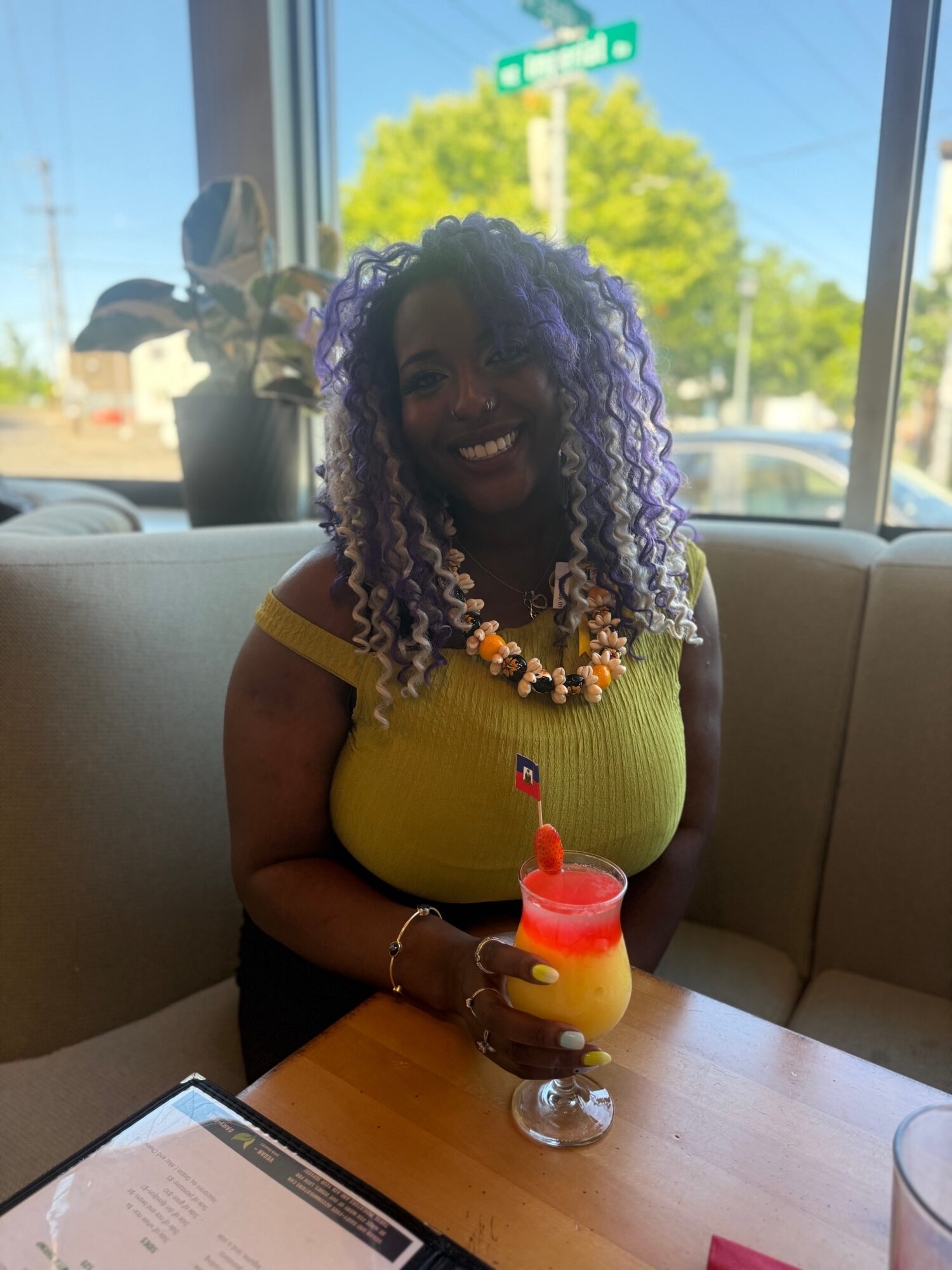 A seated person with curly purple hair wears a yellow top and holds a layered drink at a table.