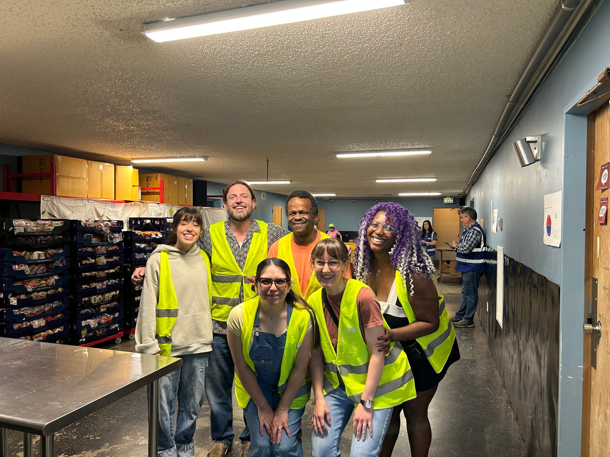 Five people wearing bright safety vests pose in a warehouse aisle lined with shelves and boxes.