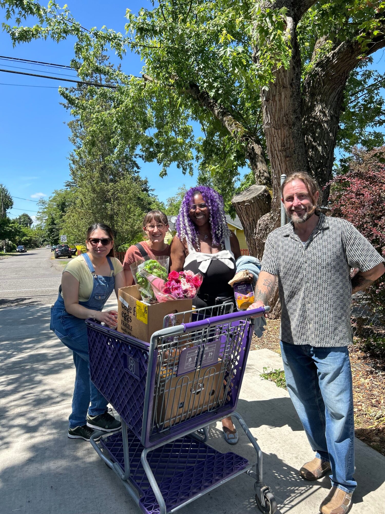 Four people stand beside a shopping cart under a tree on a sunny sidewalk.