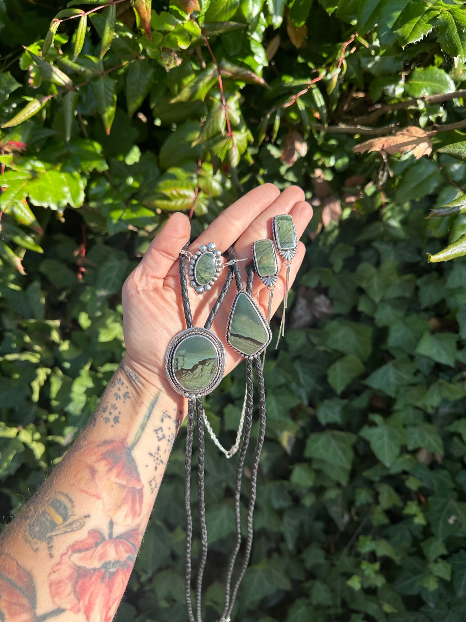 Hand with tattoos holding various rings and jewelry against green leafy background.