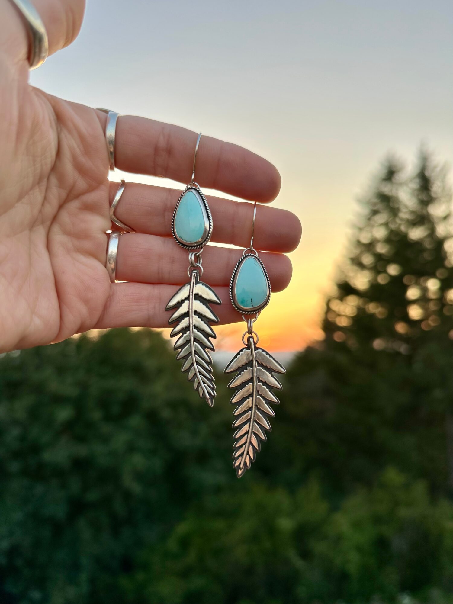 Hand holding turquoise and silver leaf earrings against a sunset sky with trees in the background.