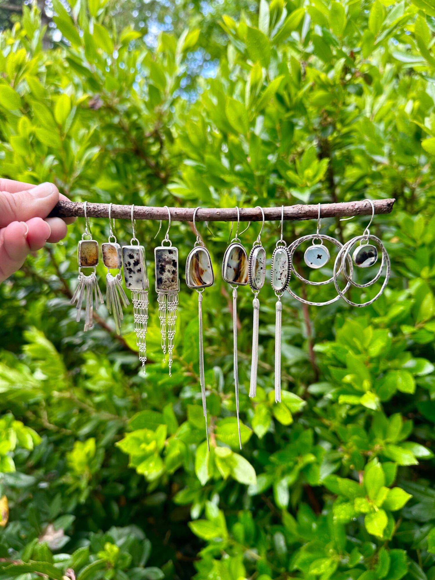 Hand holding a jewelry display with various silver earrings and necklaces, green leafy background, bright outdoor setting.