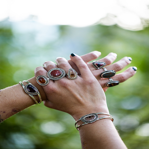 Two hands with multiple rings and bracelets, outdoors with blurred green background.