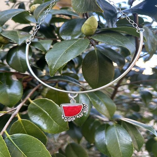 Necklace with a red semi-circle pendant hanging among green leaves.