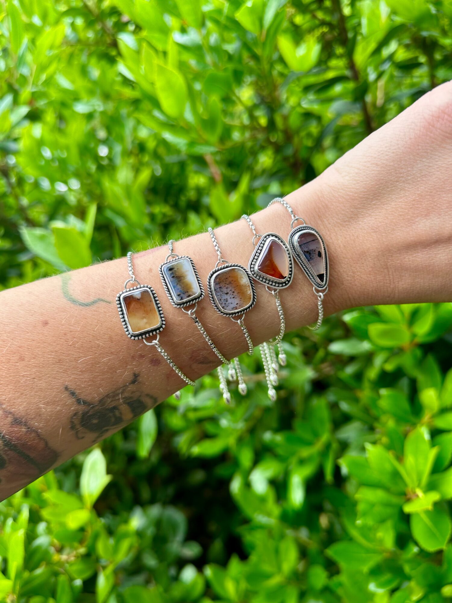Person's arm with five silver bracelets featuring rectangular and oval stones, against green leafy background.