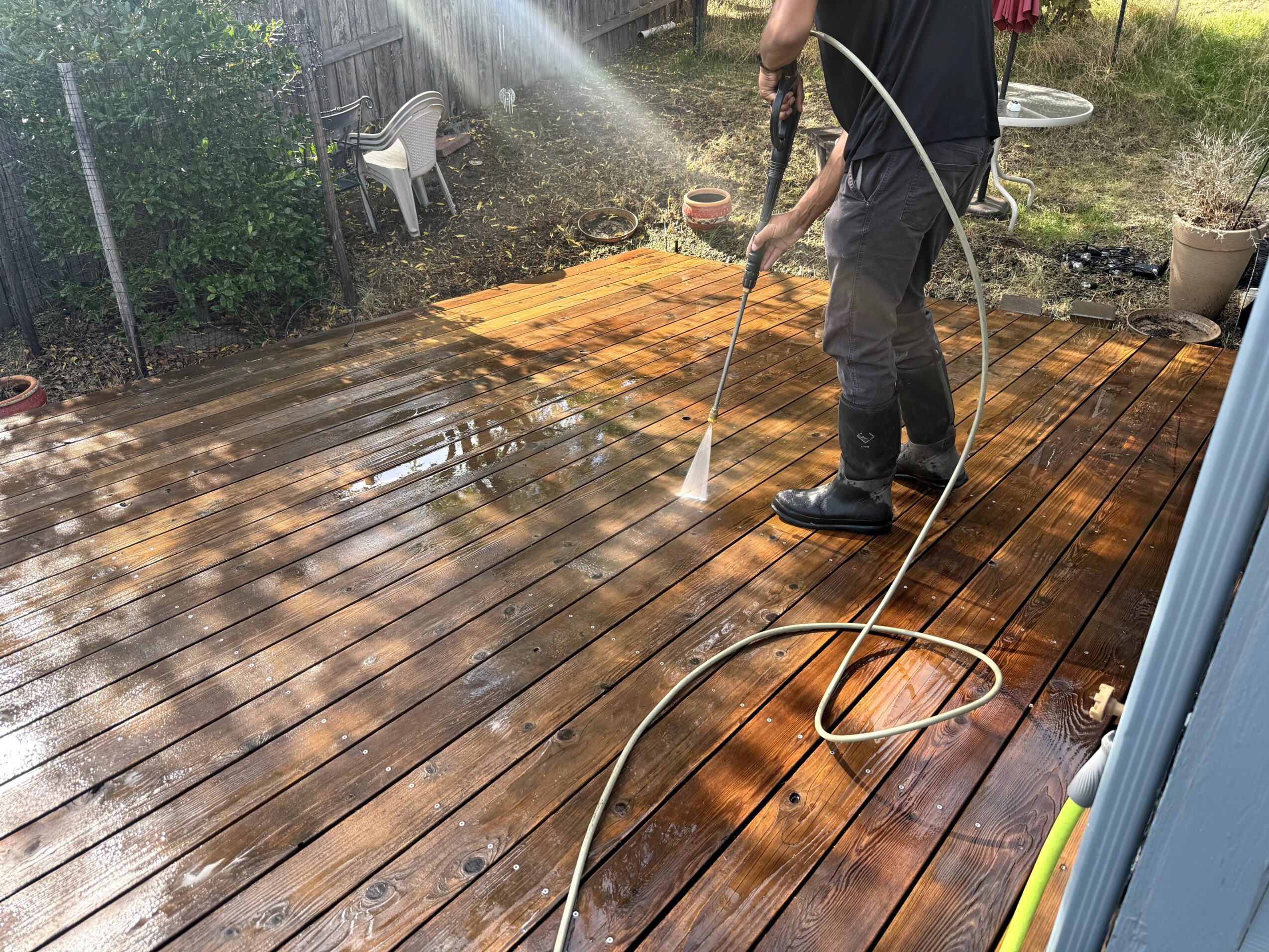 Person using a pressure washer on a wooden deck outdoors, with sunlight shining and garden elements in background.