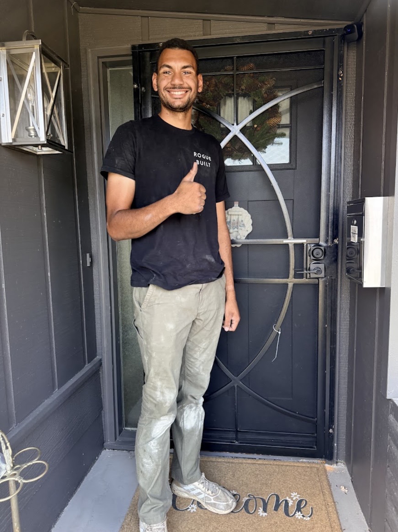 Man standing in front of a door, smiling and giving a thumbs-up, wearing a black shirt and light-colored pants.