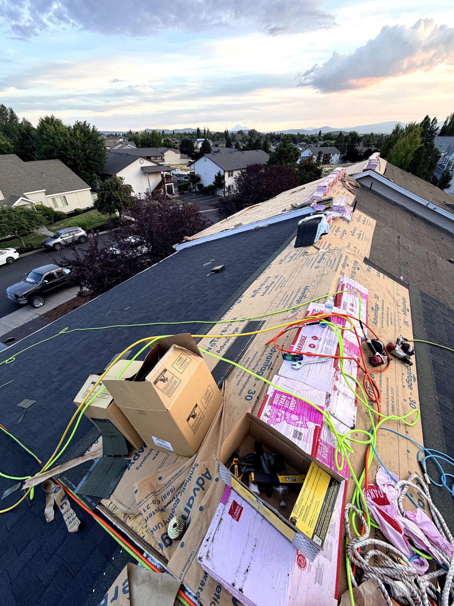 View from rooftop showing construction tools, boxes, and materials with houses and trees in the background under a partly cloudy sky.