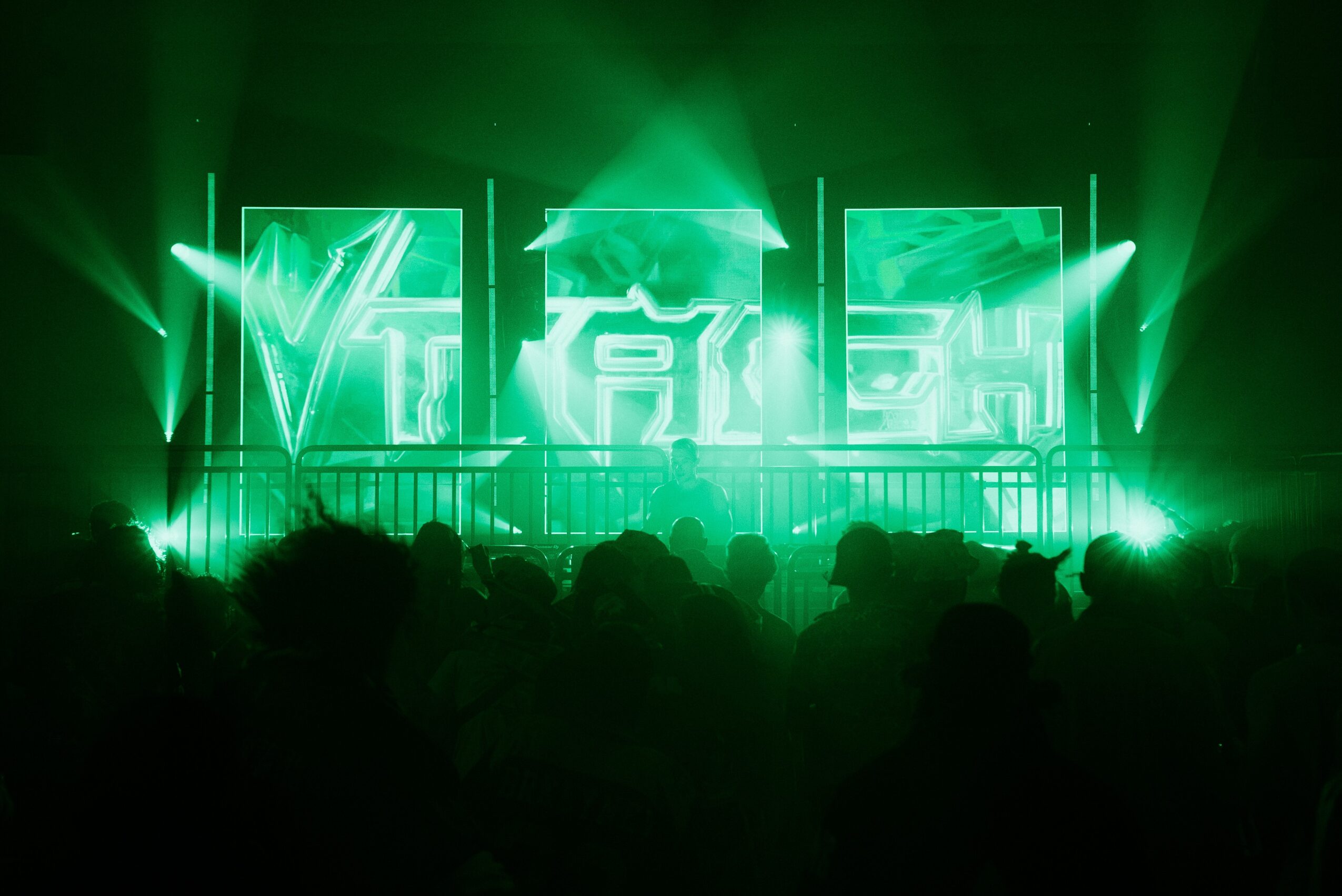 Concert stage with green lighting, screens displaying symbols, and an audience silhouette in front.
