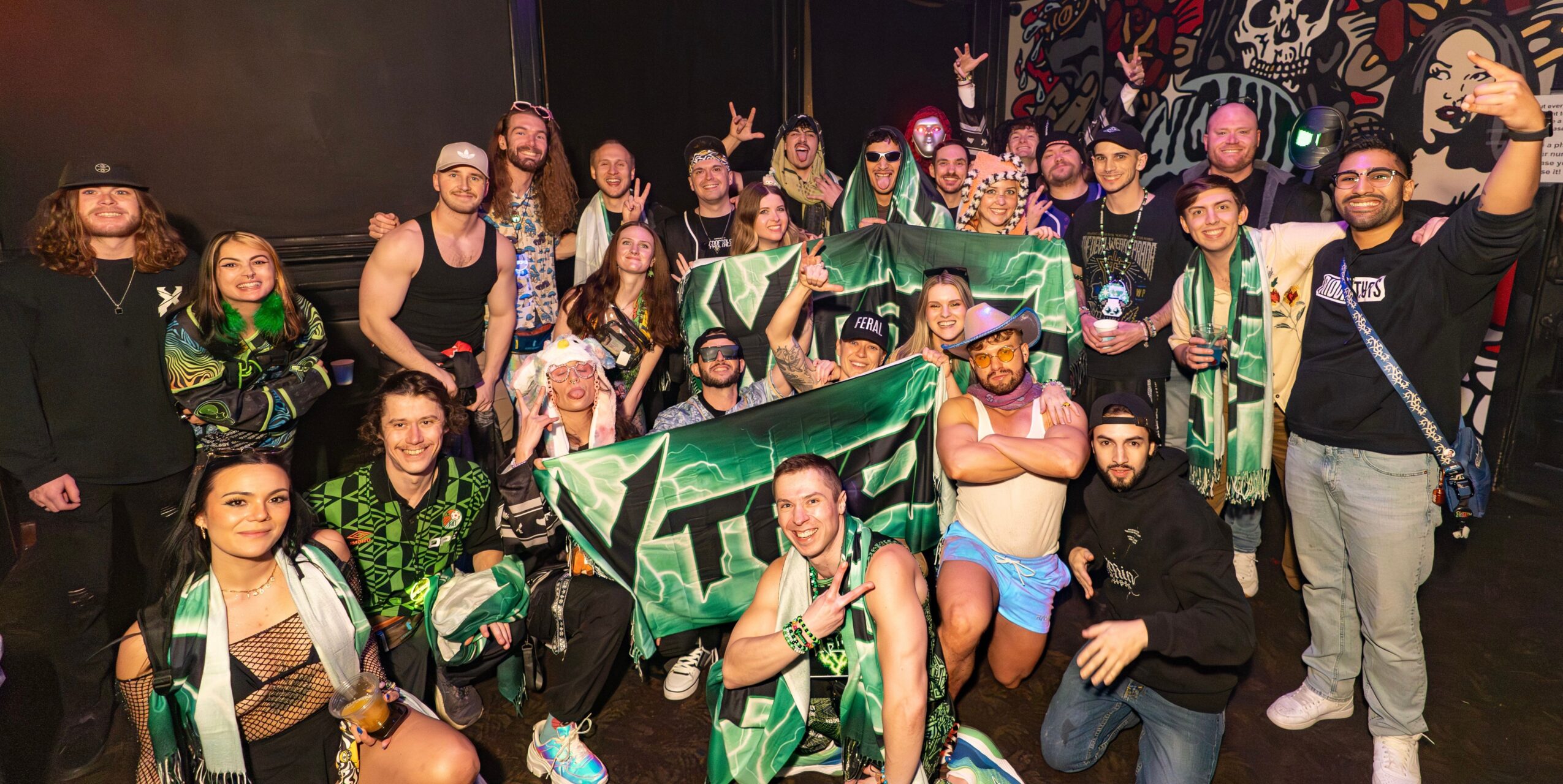Group of people celebrating, holding a green and black flag, some making peace signs, in a dark indoor setting.