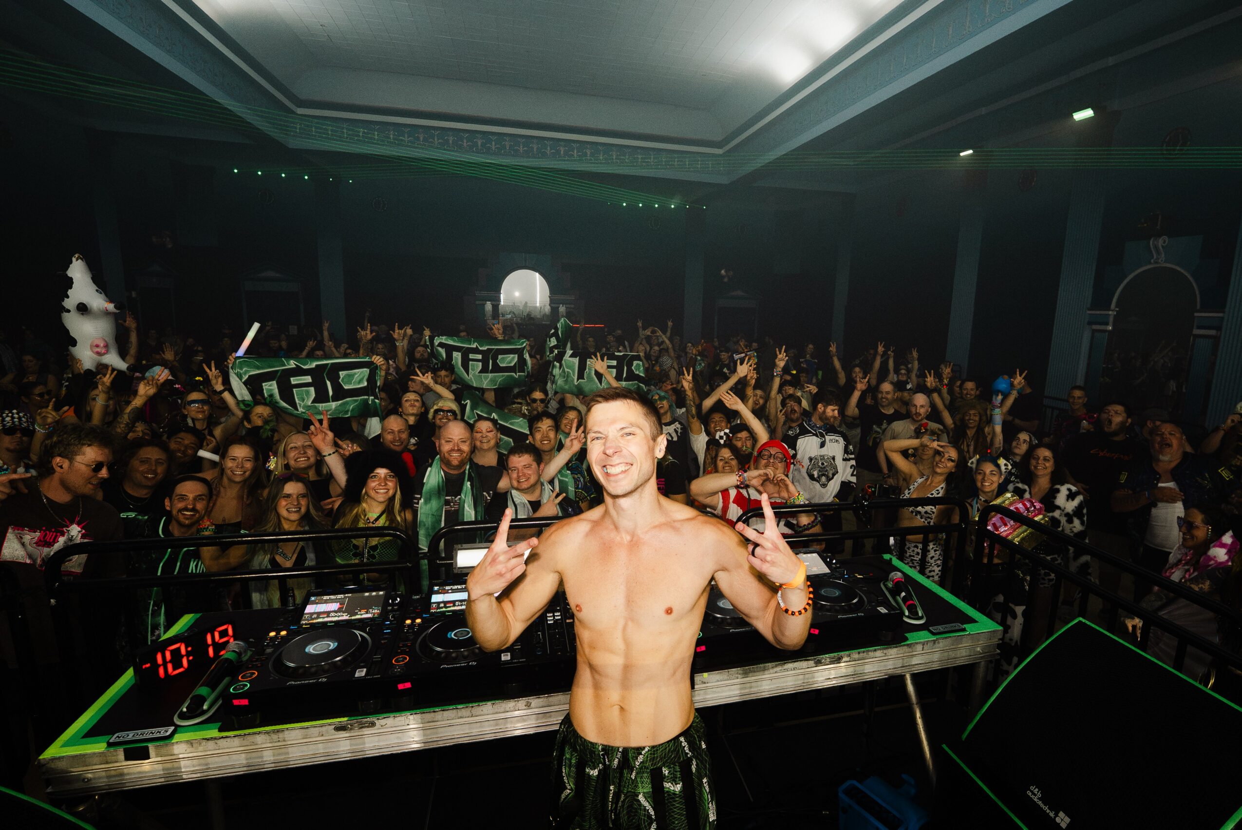 A shirtless DJ with short hair making peace signs in front of a crowd at a nightclub.