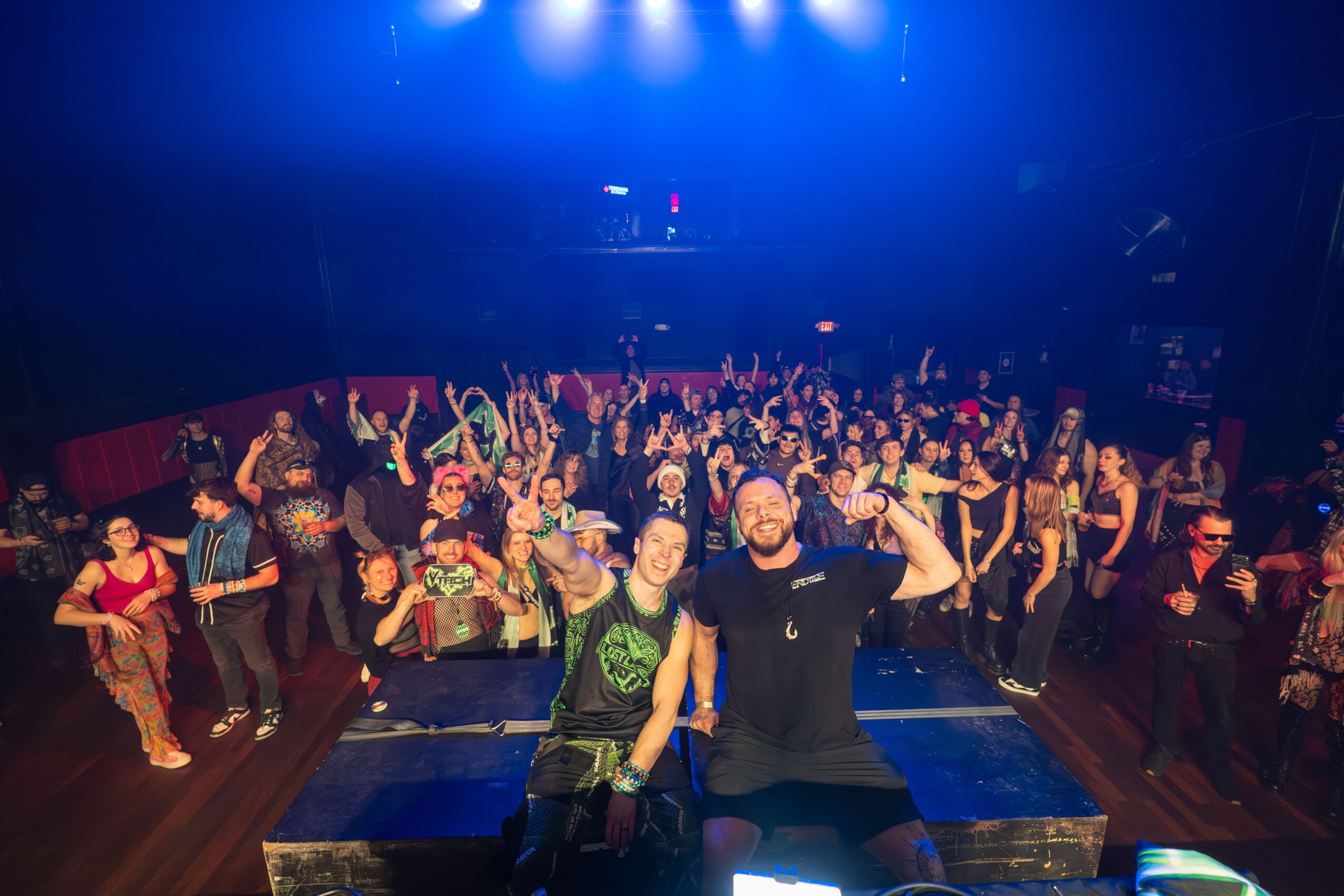 Crowd of people dancing and enjoying a party in a dark, blue-lit venue with two men in front smiling and waving.