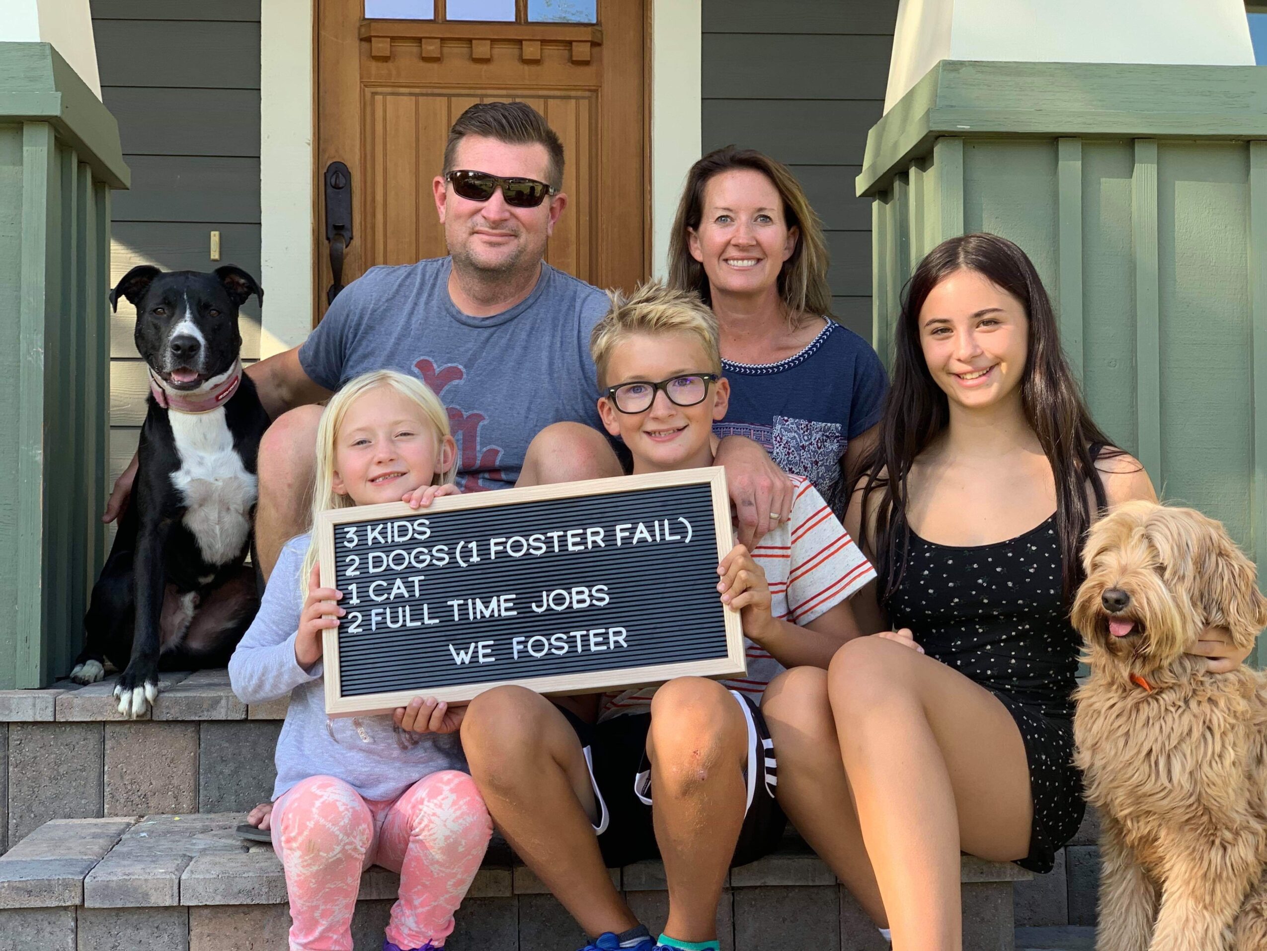 Family of six with two dogs sitting on front steps, holding a sign, smiling outside a house.