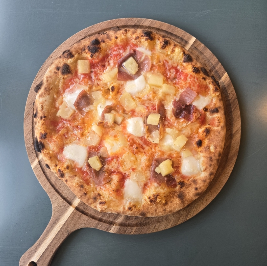 Pizza with cheese, ham, pineapple, and other toppings on a wooden serving board.