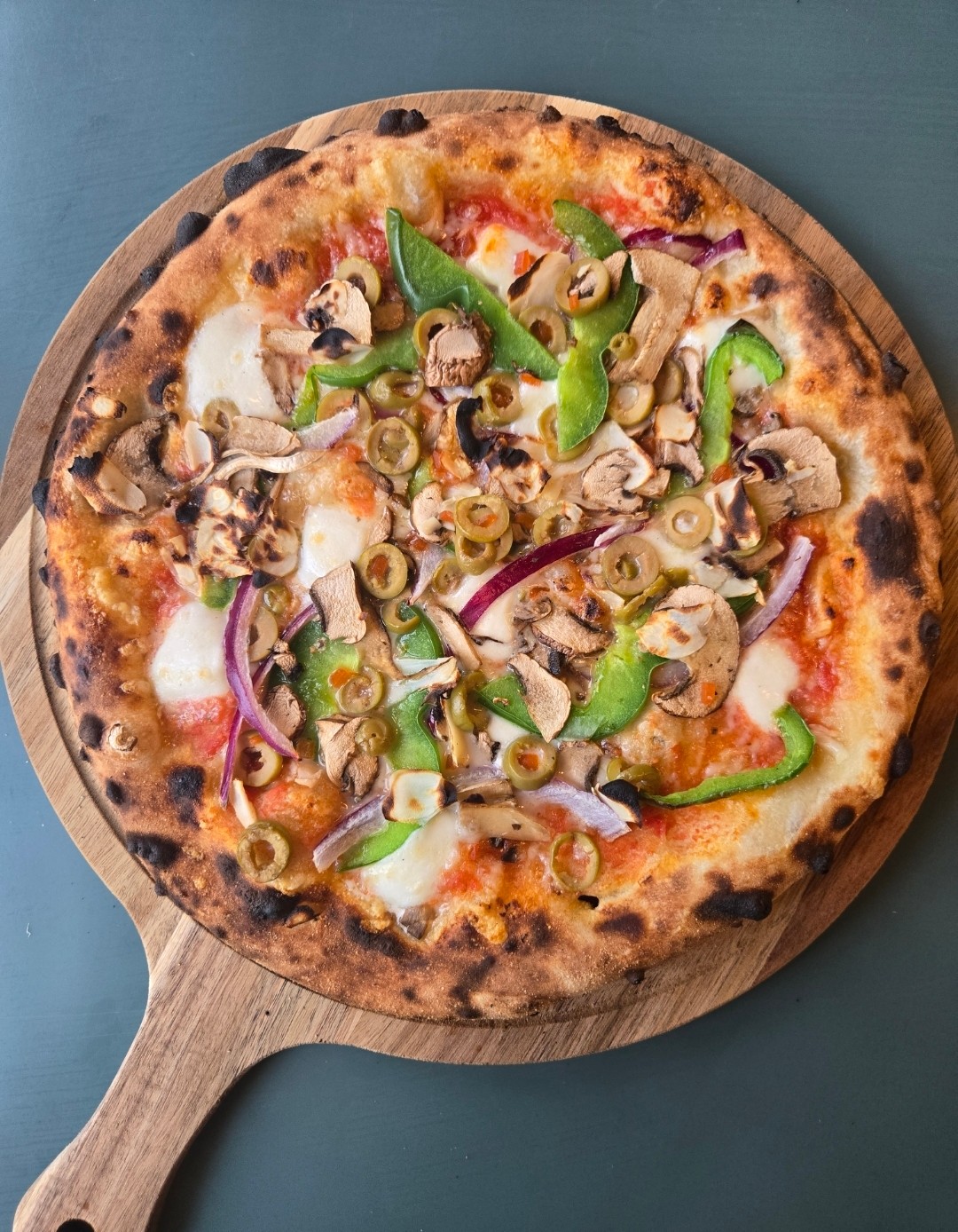 Pizza with green bell peppers, mushrooms, olives, and red onions on a wooden paddle, on a blue surface.