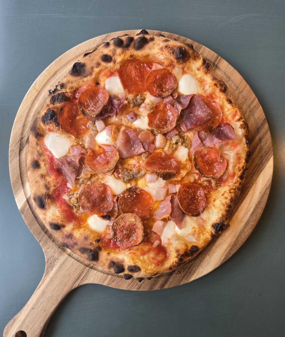 Pizza with pepperoni, ham, and cheese on a wooden serving board, viewed from above.