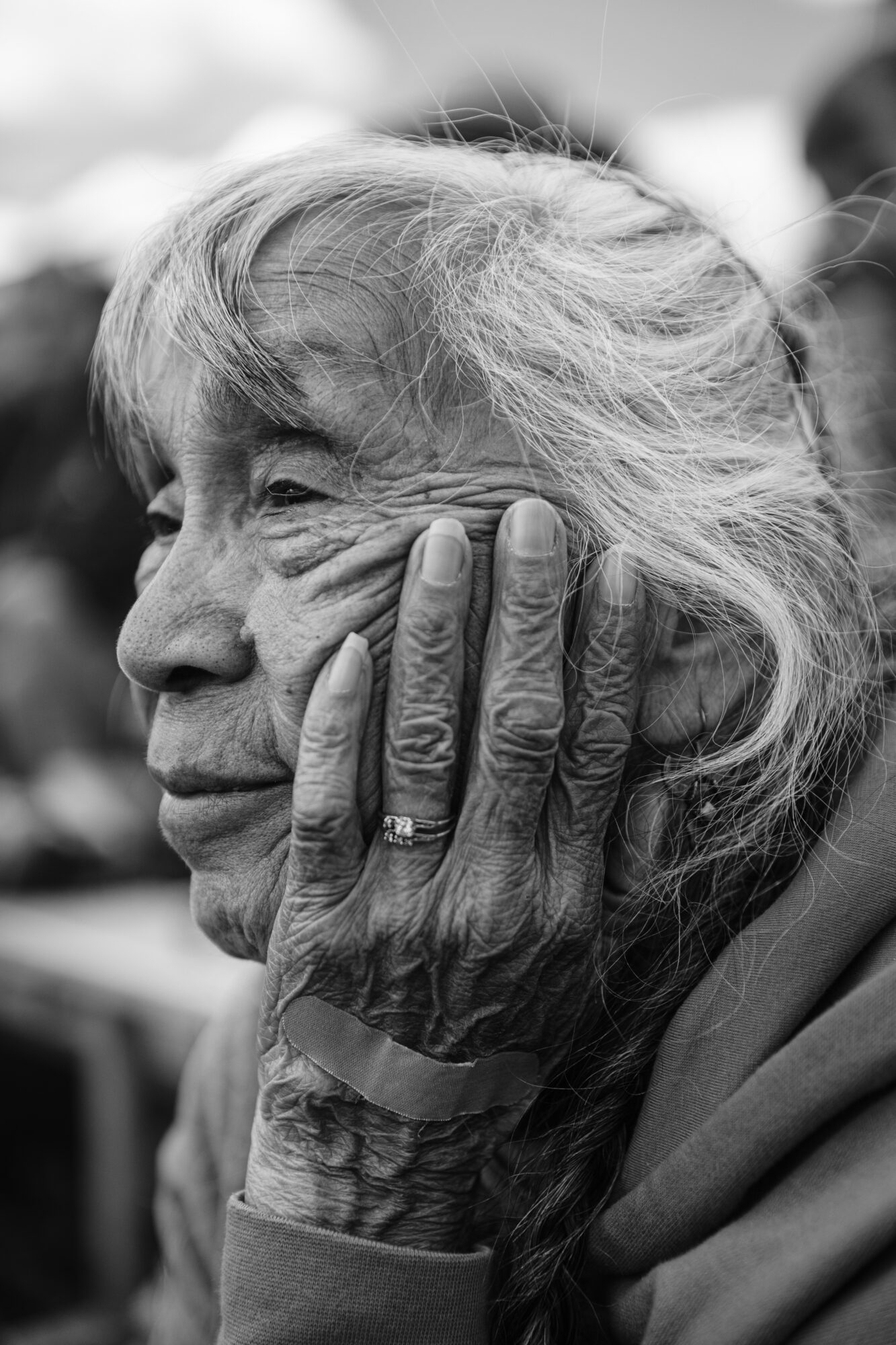 Close-up of an elderly woman with gray hair, resting her face on her hand, showing wrinkles and a ring on her finger.
